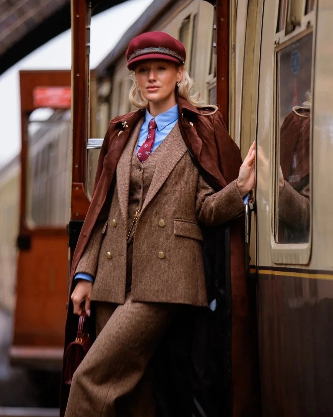 Woman in vintage train conductor uniform standing at train door, wearing a red cap, brown checked suit, blue shirt, and a patterned tie.