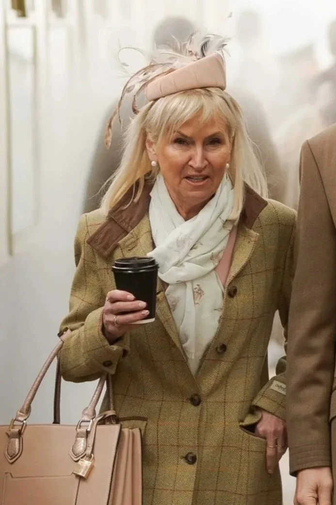 A woman with blonde hair wearing a plaid coat, scarf, and hat with feathers, holding a disposable coffee cup, standing indoors.