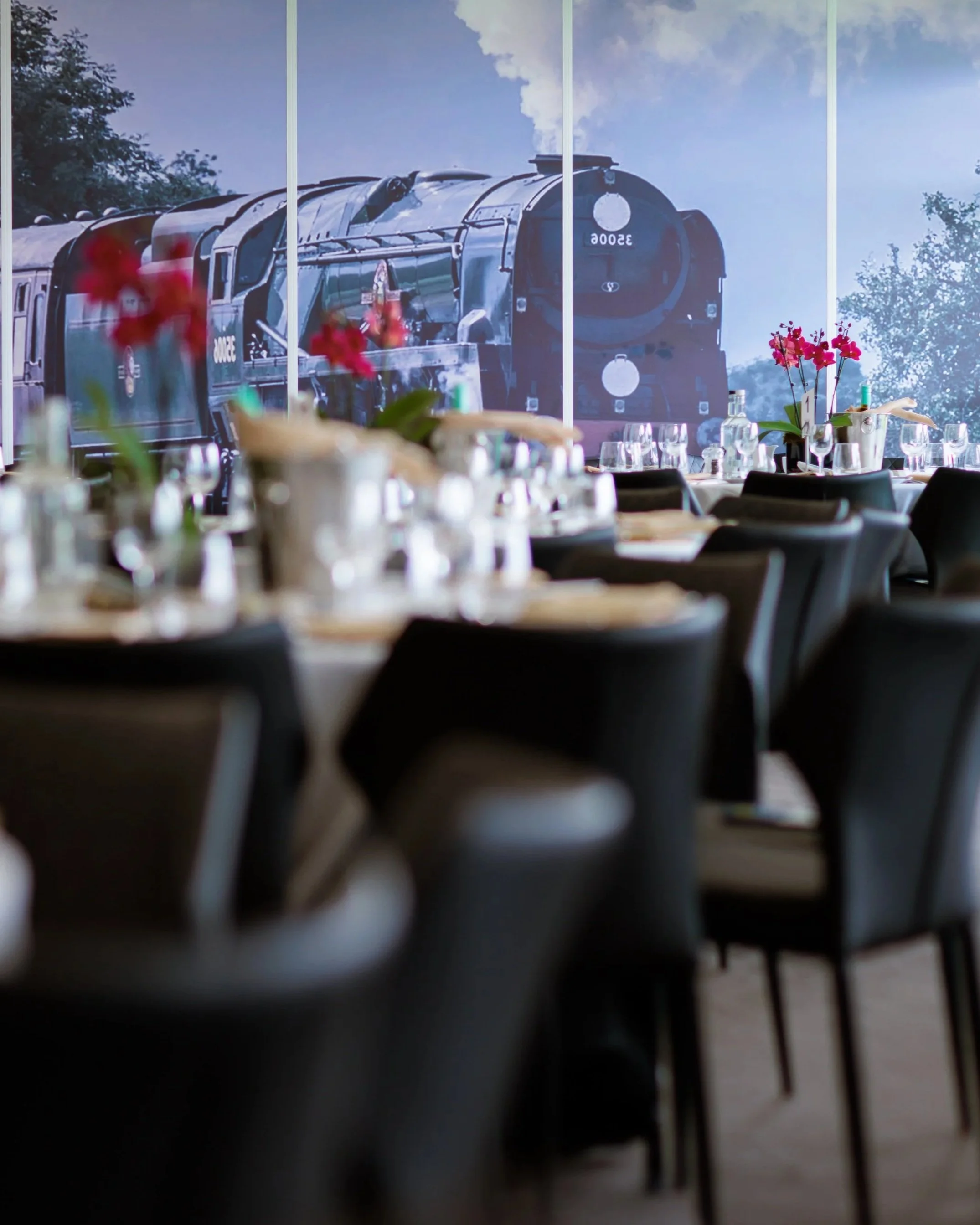 A restaurant dining area with tables set with cutlery and glasses, and a large window showing a steam locomotive outside.