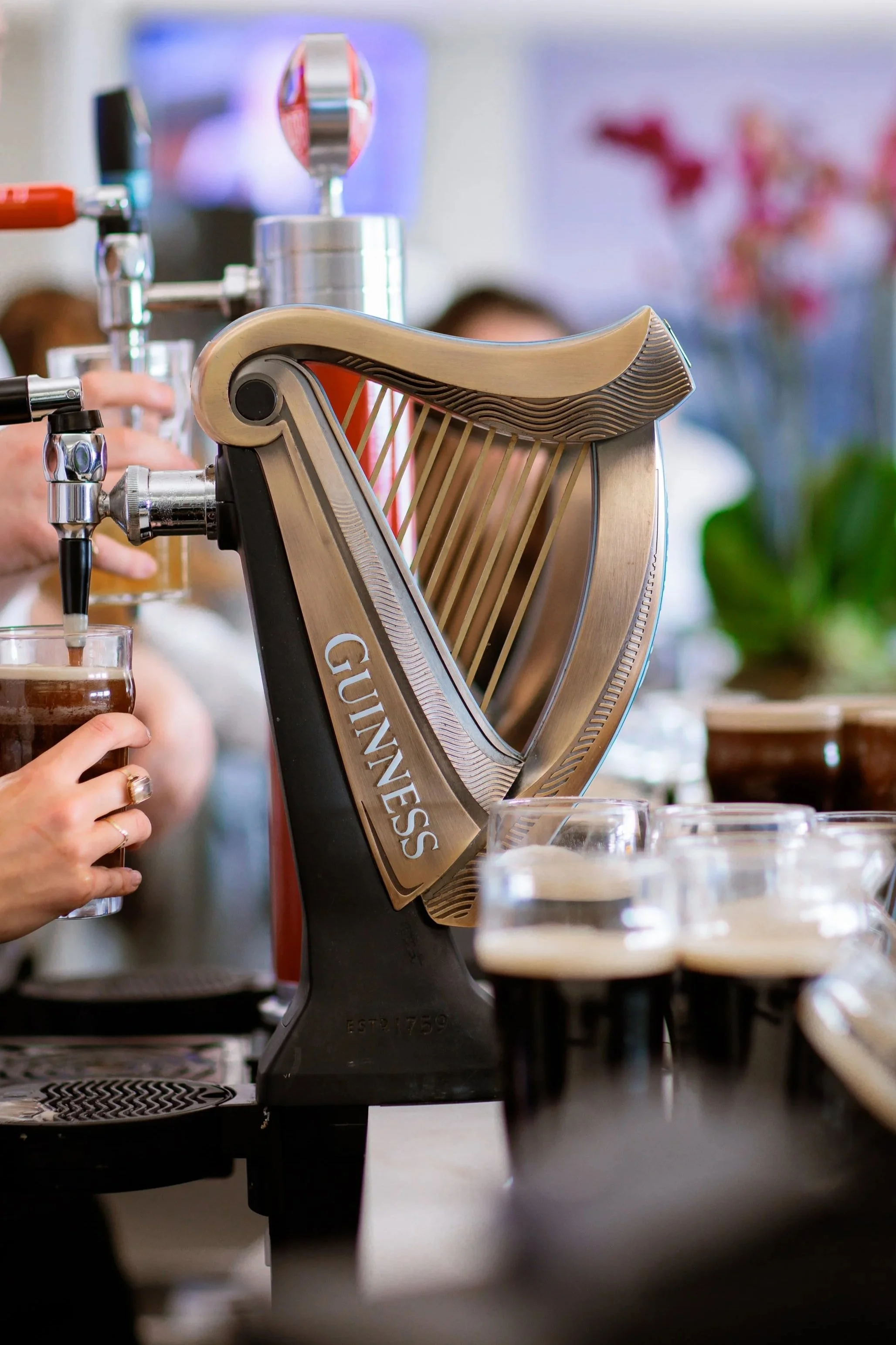 A Guinness beer tap with glasses of dark beer with foam on top on a bar counter, in a lively pub setting.