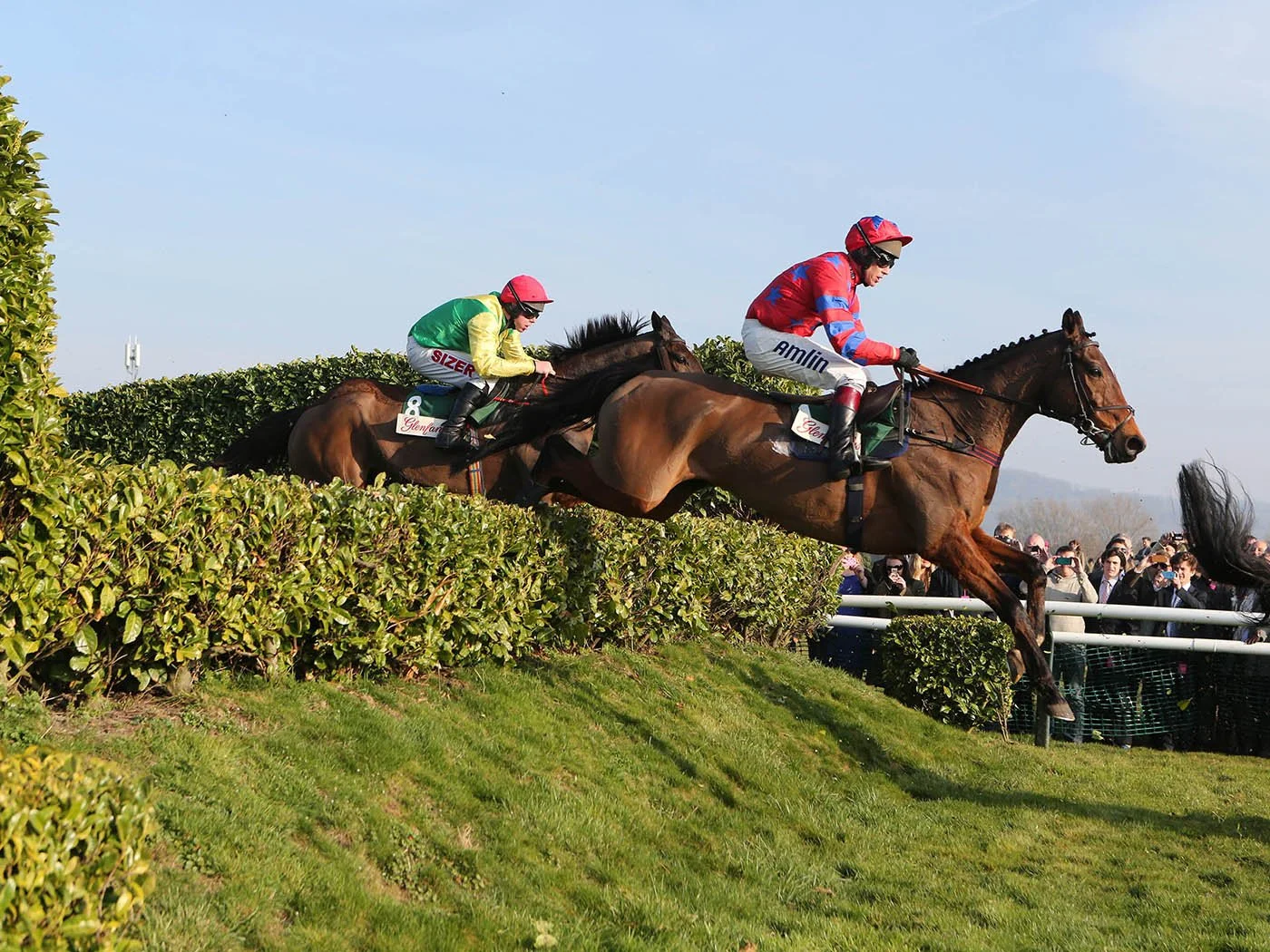 Horses and jockeys jumping a fence during a horse race, with spectators watching in the background.