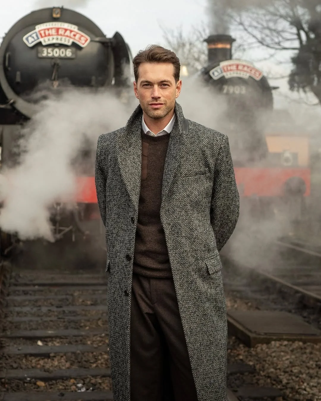 A man stands on train tracks in front of an old steam train emitting steam, wearing a gray herringbone coat and a white shirt.