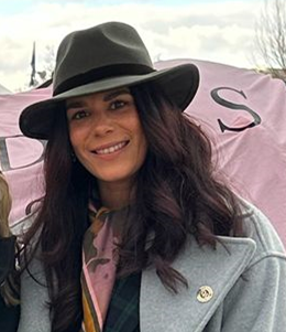 Woman smiling, wearing a gray coat and black hat, outside with a pink flag or banner in the background.