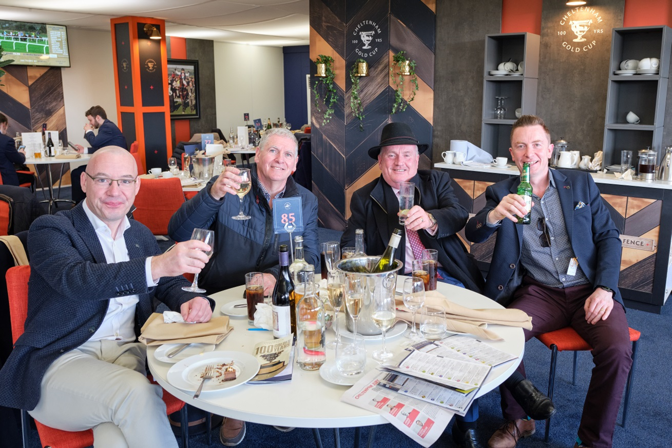 Four men sitting at a round table in a restaurant celebrating with drinks, bottles and glasses of wine, and partially eaten desserts, with a TV and other diners in the background.