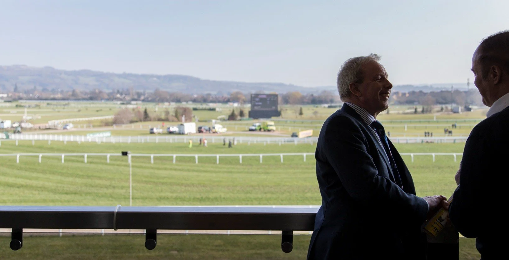 Two men in suits shaking hands and smiling inside a viewing box at a racecourse, with a green racetrack and distant hills in the background.