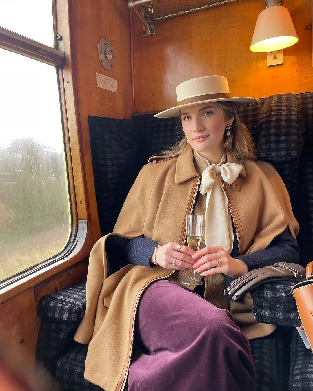 A young woman with wavy hair, wearing a beige wide-brimmed hat and vintage style clothing, sitting inside a train carriage with wood paneling, holding a glass of champagne.