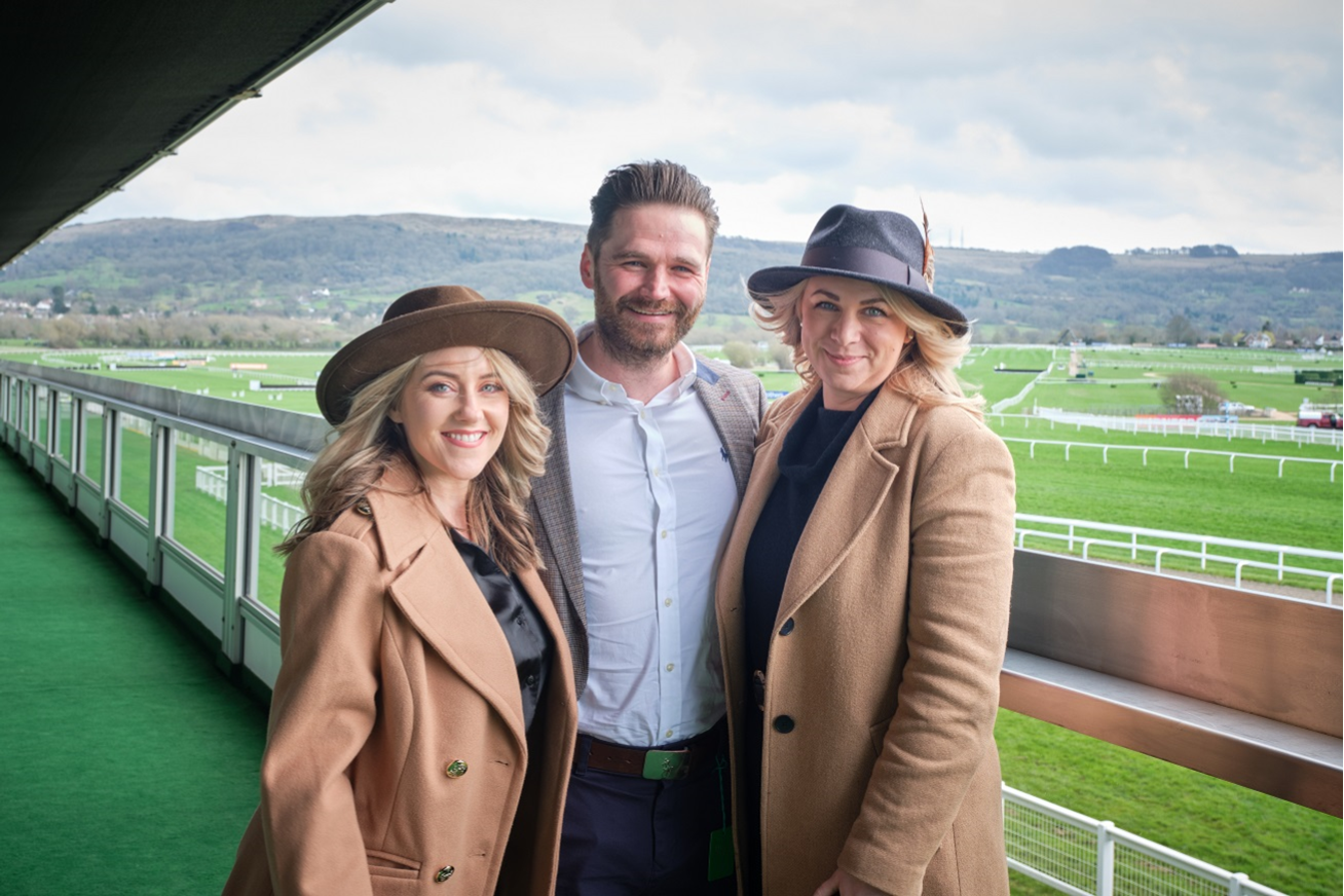 Three people standing on a balcony overlooking a horse racing track on a cloudy day.