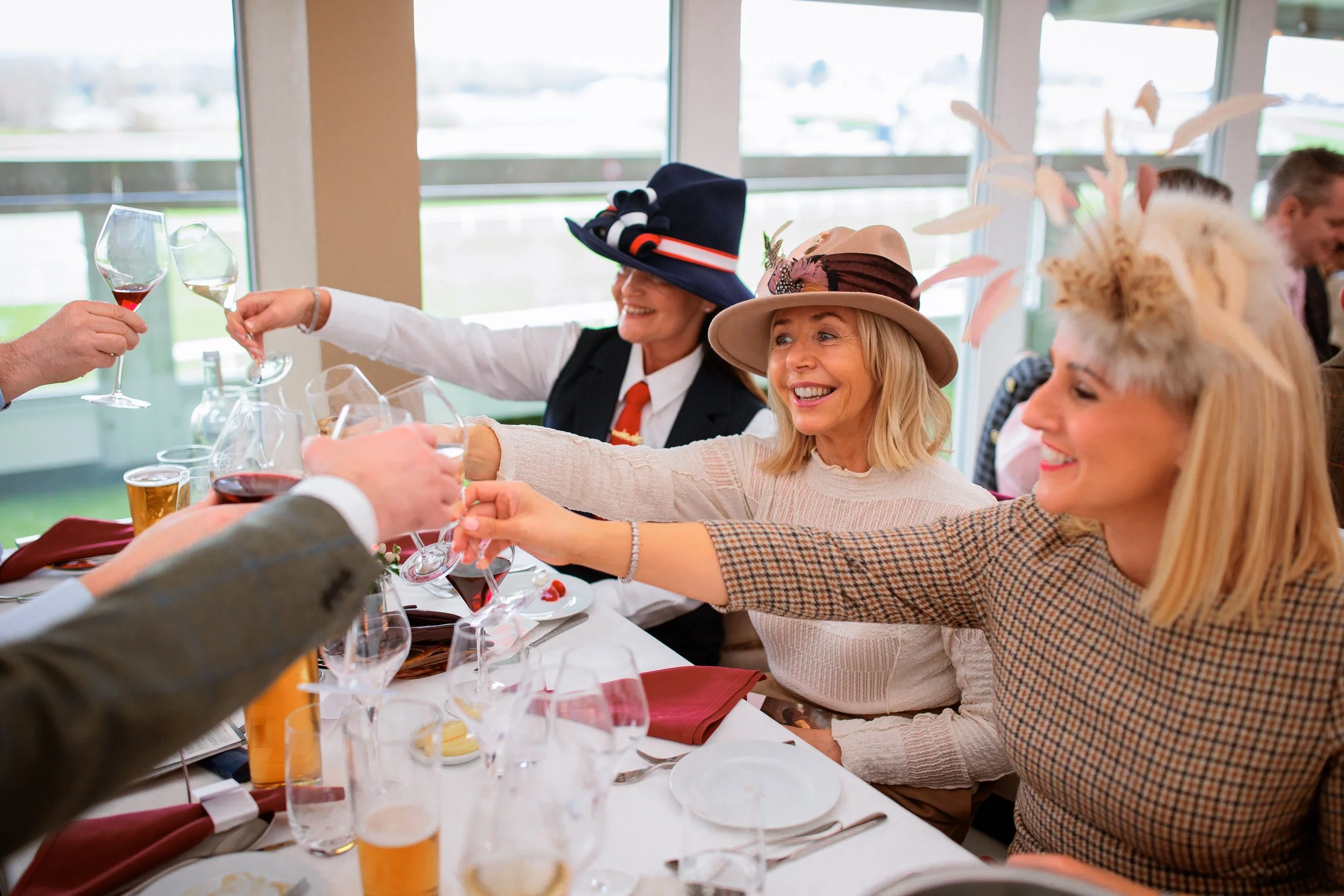 Celebrating at a social gathering with women wearing fancy hats, toasting glasses, and smiling around a table with drinks and plates.
