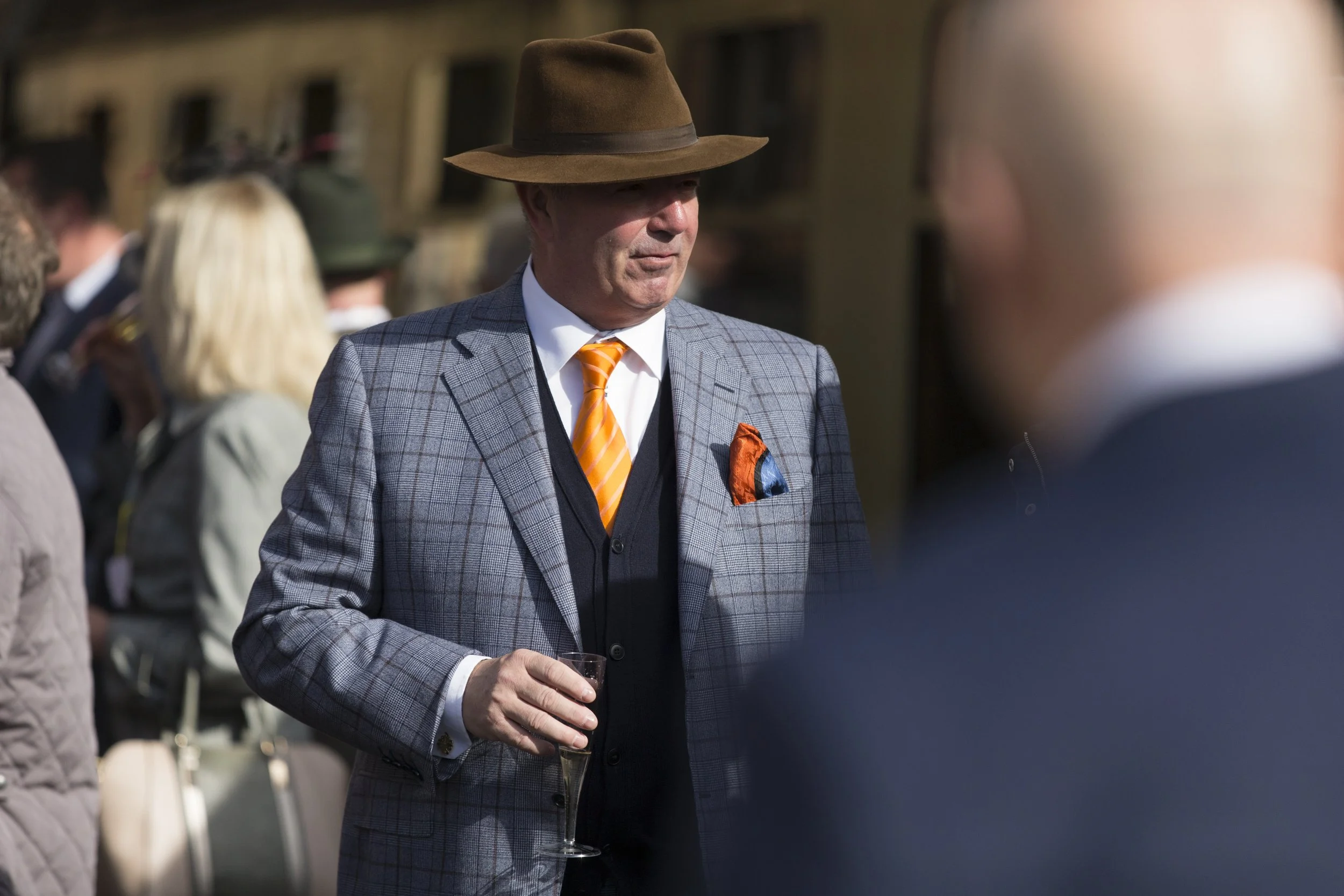 A man in a gray plaid suit with an orange tie and pocket square, wearing a brown fedora, holding a glass of wine at an outdoor event.