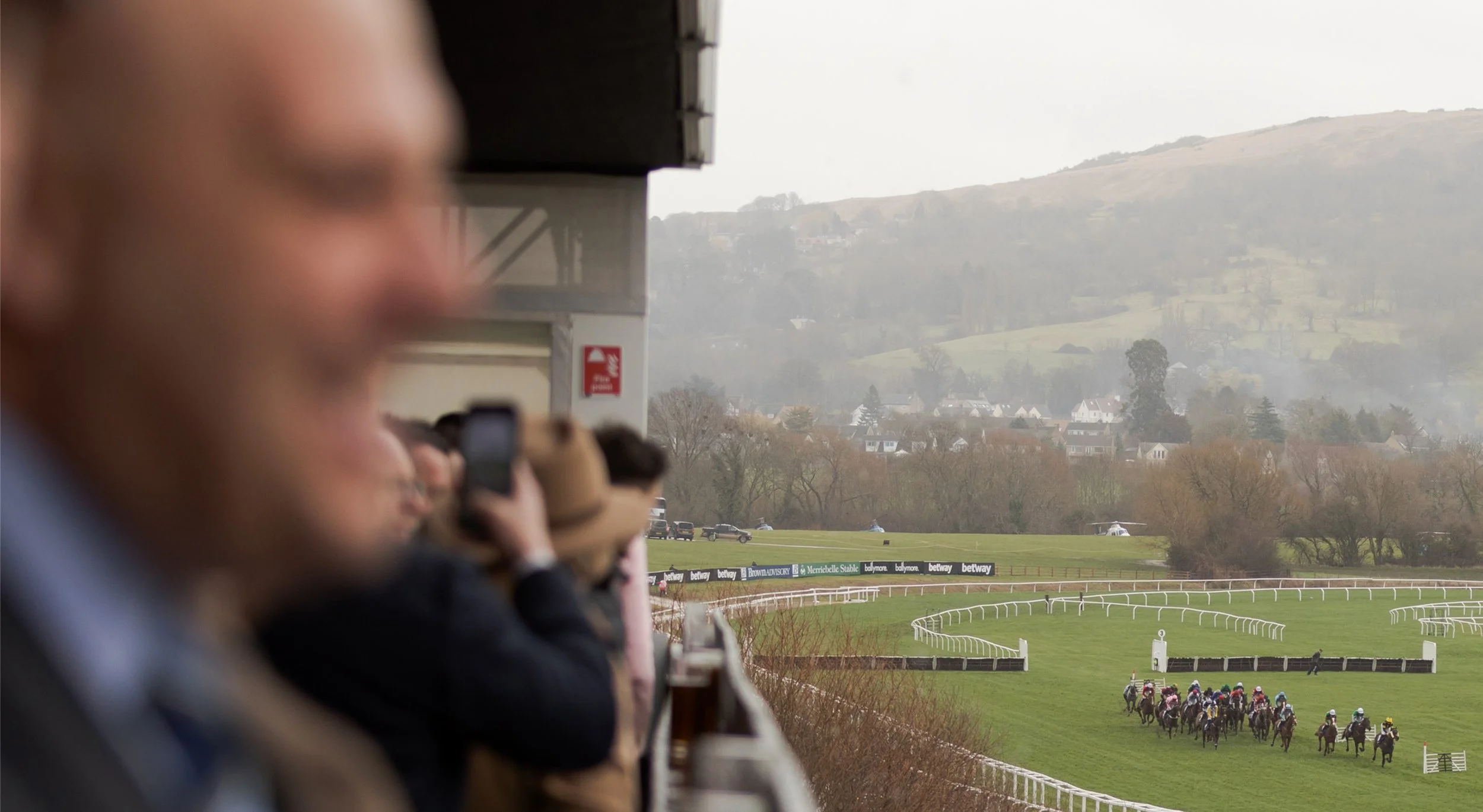 Horse race at a racetrack with spectators watching from the stands, scenic hillside in the background.