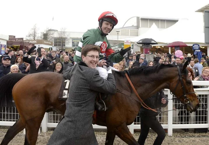 Jockey on horseback celebrates with a trainer as a crowd watches at a horse racing event.