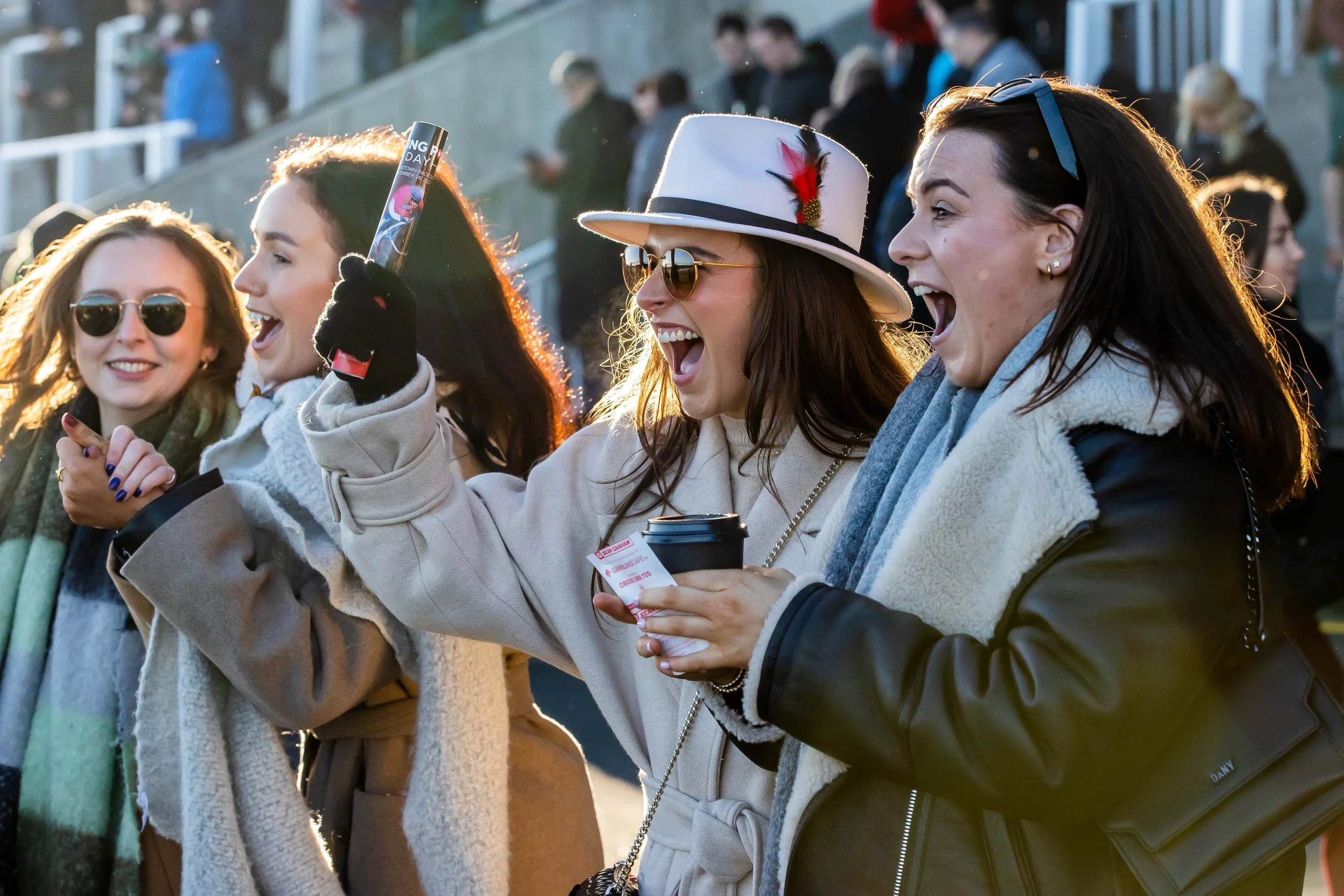 Four excited women at an outdoor event, wearing coats and scarves, one with a white hat and sunglasses, holding beverages and tickets, with a crowd in the background.