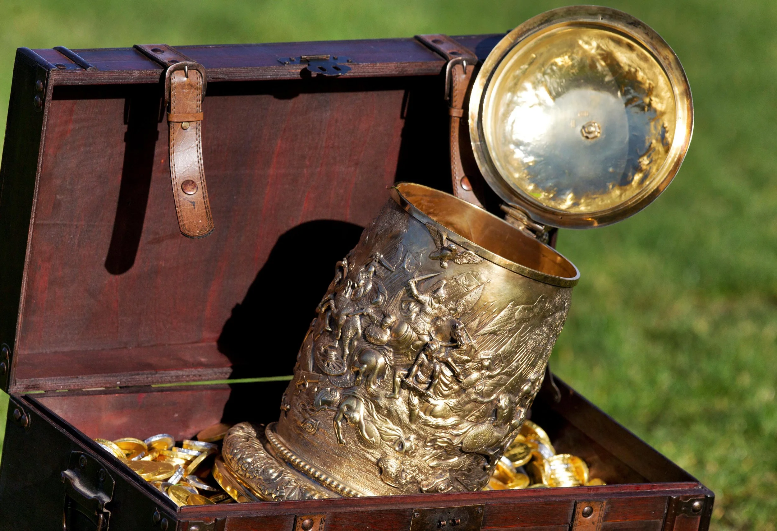 Open wooden treasure chest containing gold coins and an intricately engraved gold tankard with a hinged lid, placed on a grassy background.