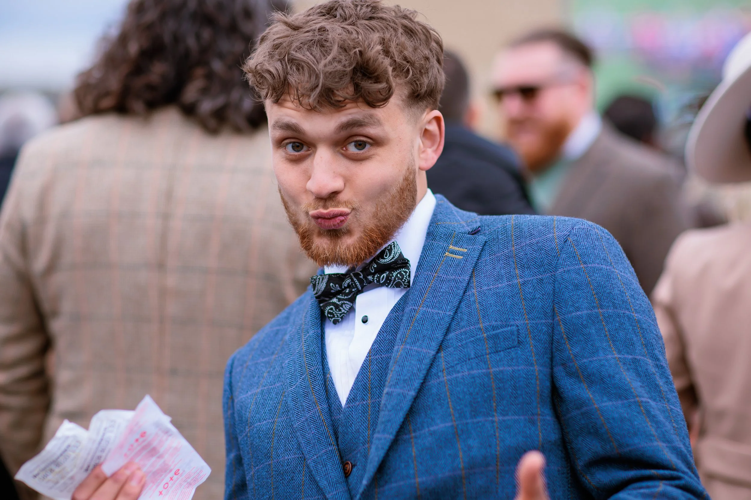 A young man with curly brown hair and a beard making a playful face at the camera. He is dressed in a blue plaid suit, white shirt, and a black patterned bow tie. He is holding a crumpled receipt in one hand, with a crowd of people around him, some w
