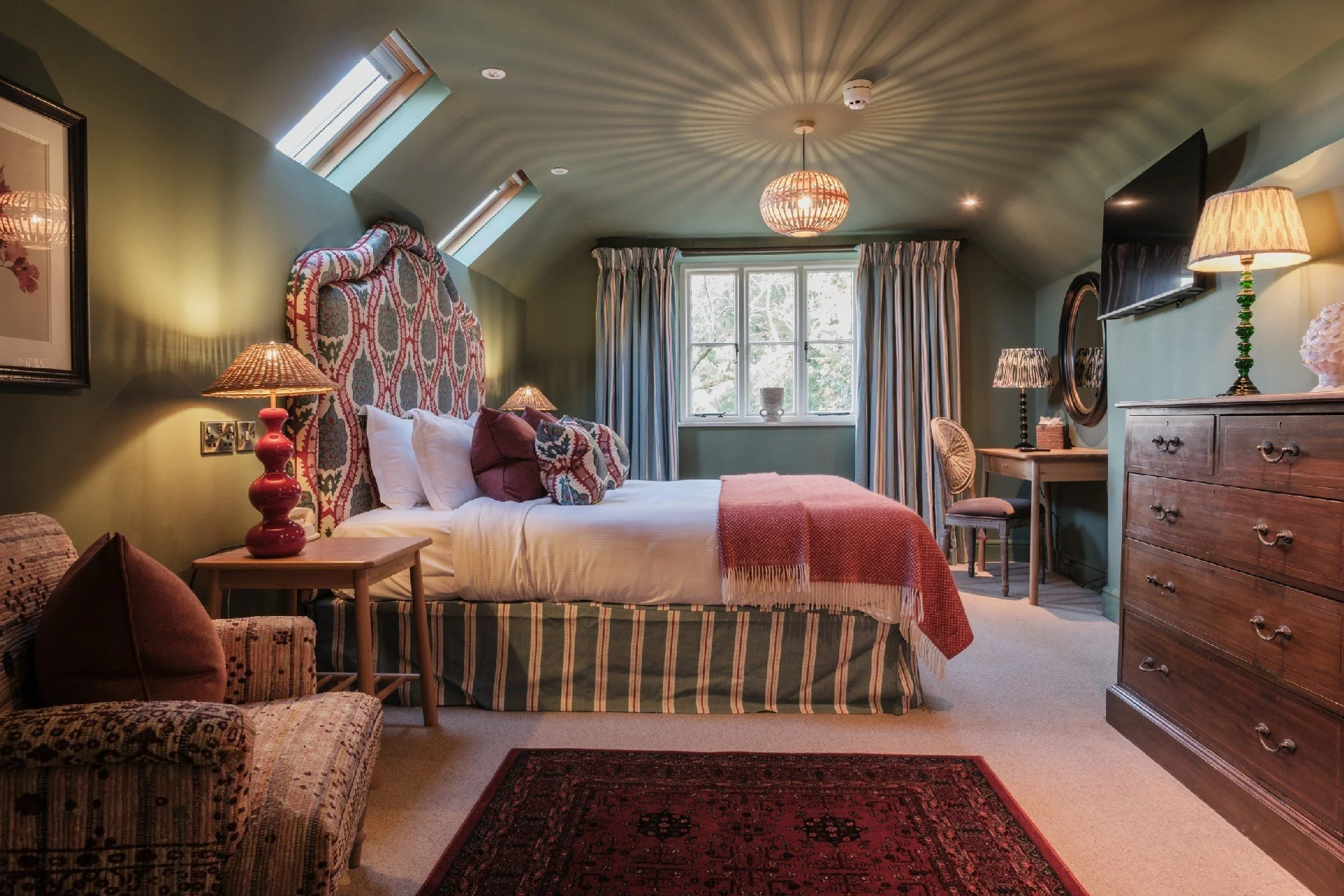 Cozy attic bedroom with patterned upholstered bed, pink throw blanket, skylights, lamps, dresser, desk, and armchair
