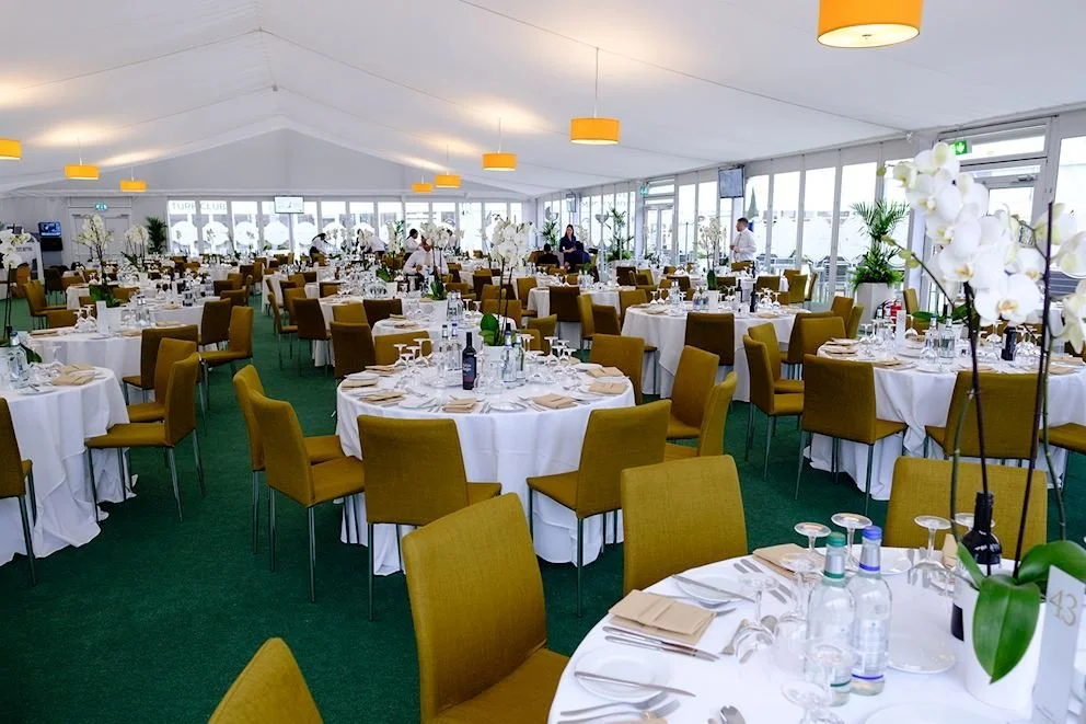 Interior of a banquet hall with round tables covered in white tablecloths, set with glassware, plates, and utensils, with yellow chairs and large windows letting in natural light.