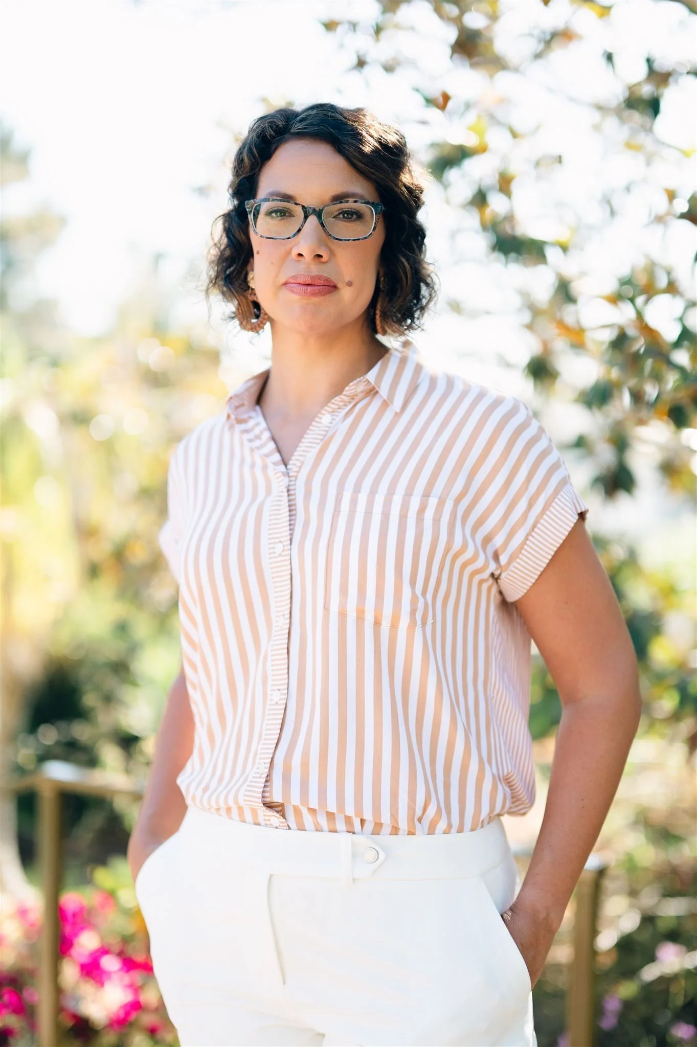 A woman therapist wearing a beige and white striped short-sleeve blouse and white pants standing outdoors with a blurred garden background. Facial expression is professional and reflective and exudes depth, strength, wisdom, and expertise.