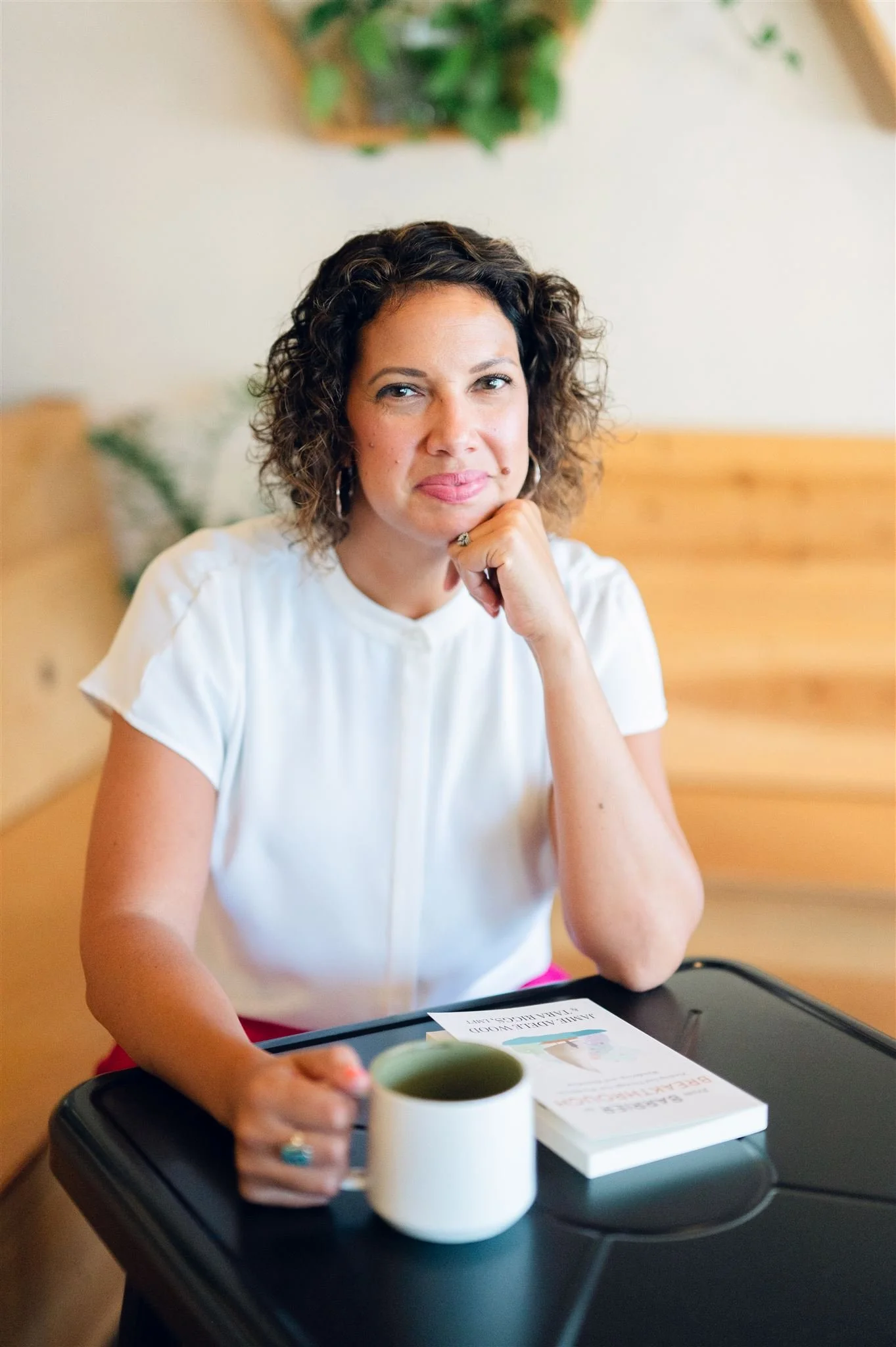 Woman with curly brown hair sitting at a desk with a white shirt, holding a coffee mug, with a book and paper on the desk. Woman appears easy to connect to, warm, humourous, and kind, inviting relationship, belonging, and authenticity.