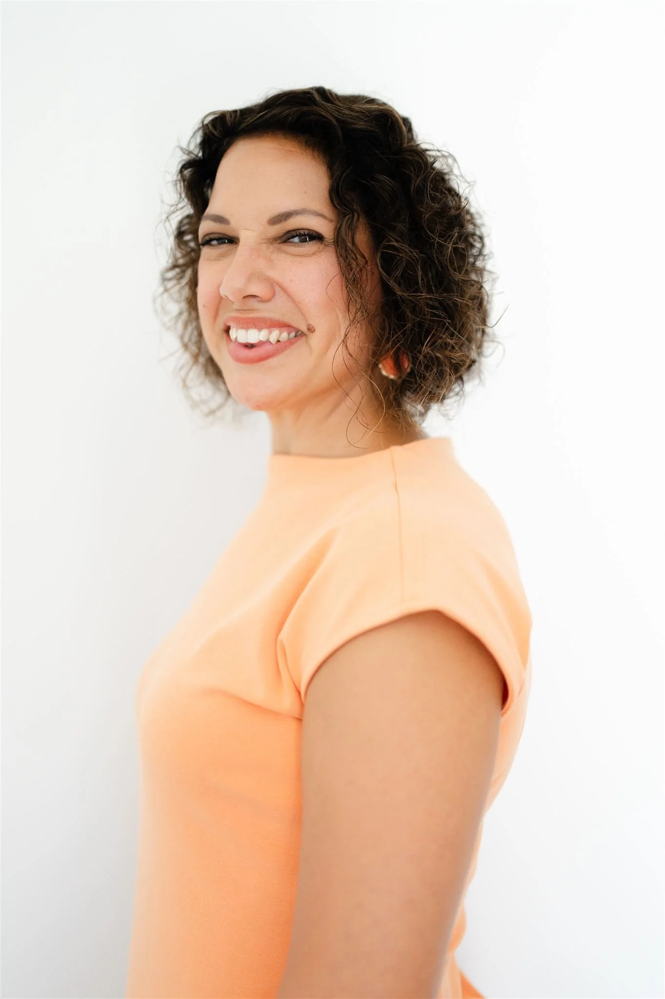 A woman with short, curly brown hair smiling and looking over her shoulder, wearing a peach-colored top against a plain white background.