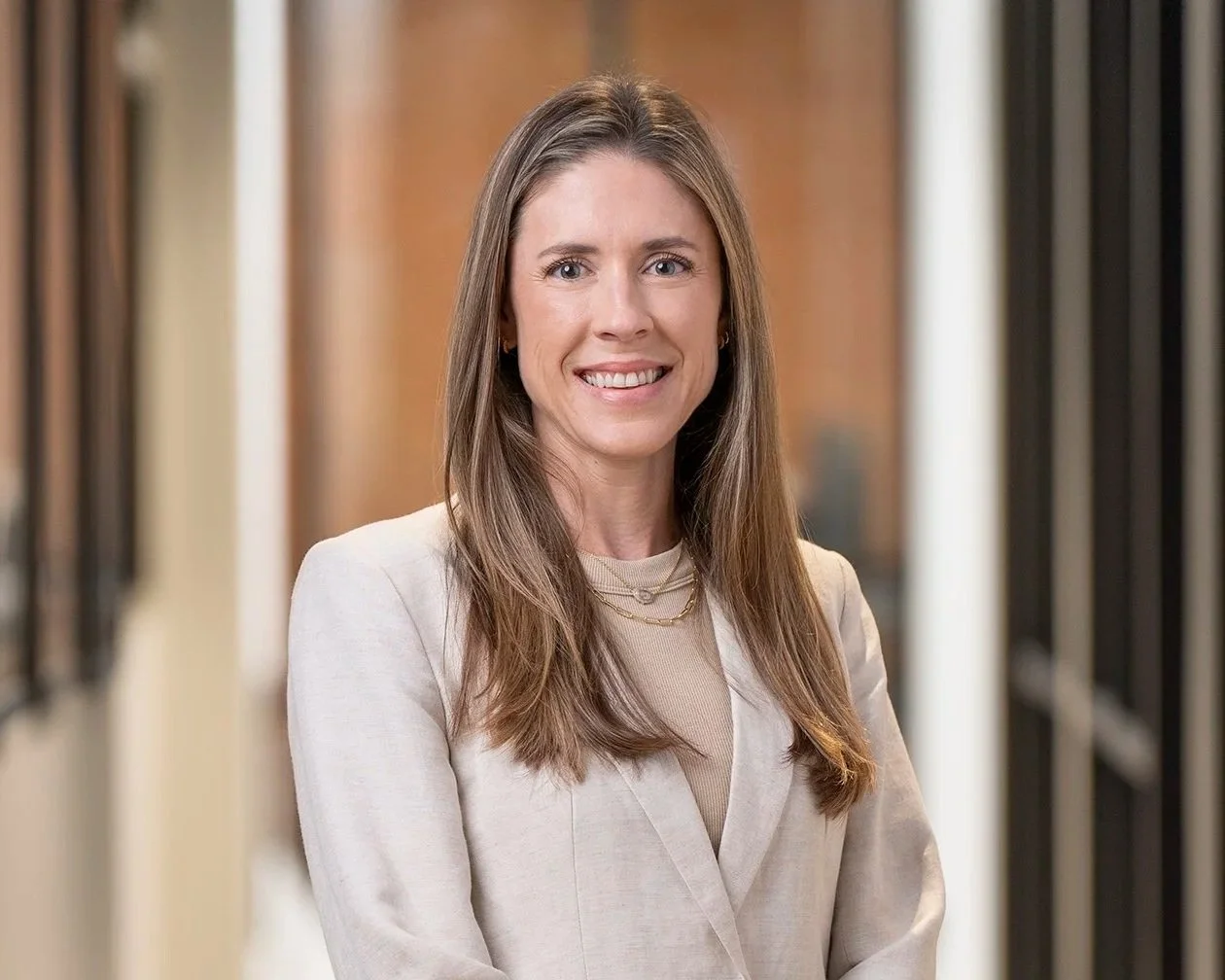 A woman with long brown hair wearing a light-colored blazer, smiling and standing in a hallway.