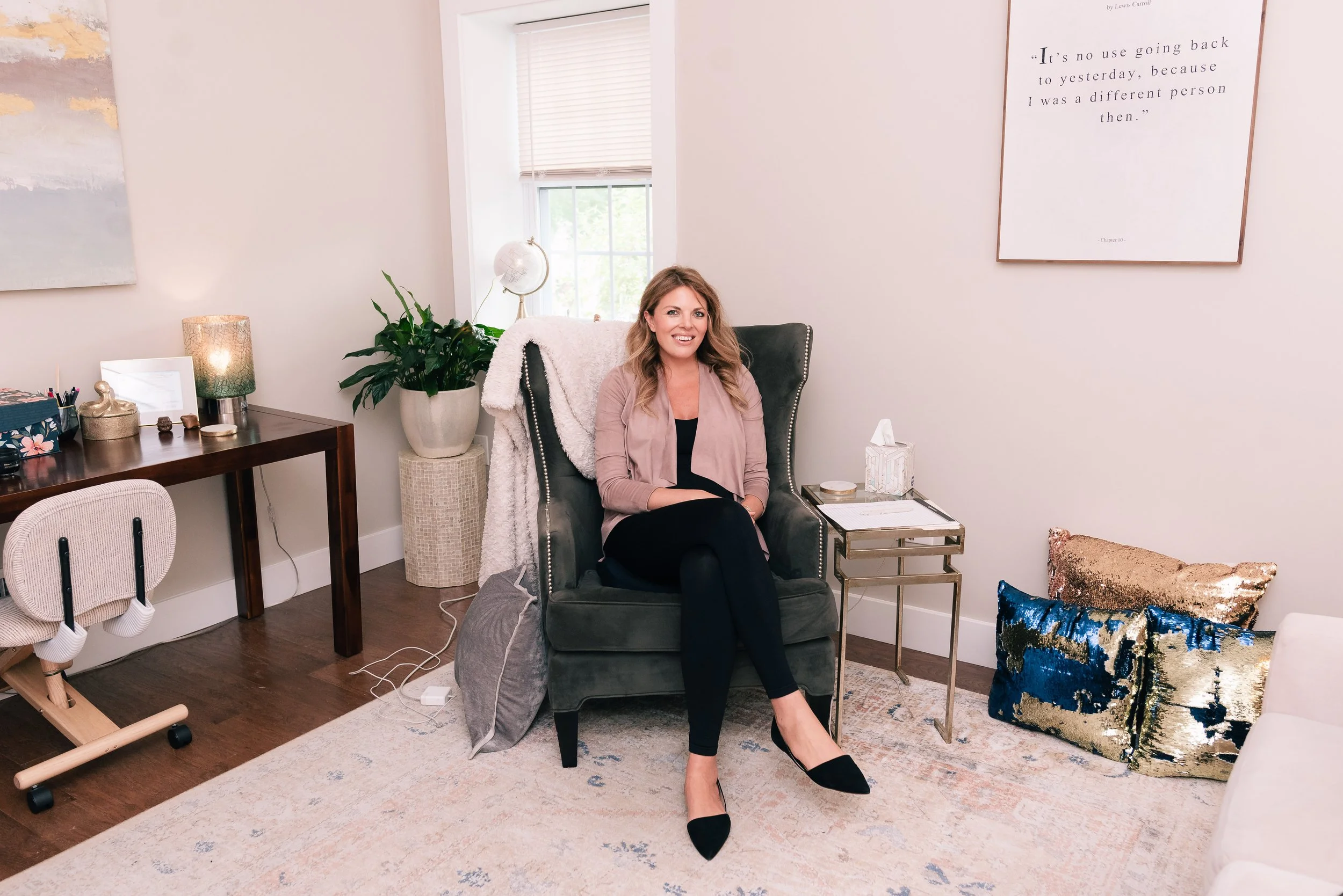 A woman sitting on a gray armchair in a living room, smiling at the camera. The room has decorated walls, a large window with blinds, a side table with decorative items, a big potted plant, and a coffee table with tissues and books. There are colorful pillows on the floor and a patterned area rug.