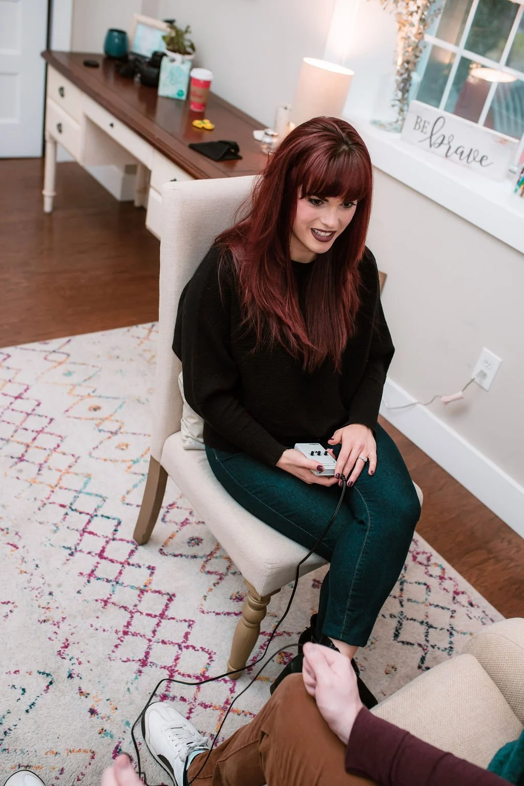 A woman with red hair and dark lipstick sitting on a white chair in a therapy room, holding EMDR tappers with a male client sitting in front of her.