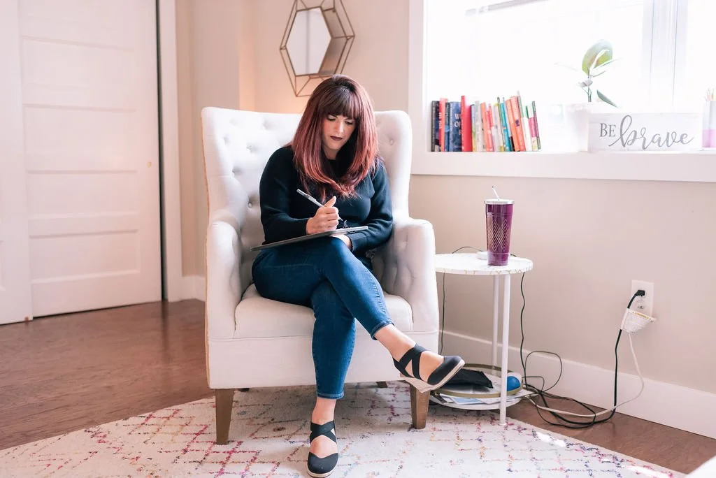 A woman with shoulder-length reddish-brown hair sitting on a white armchair, writing in a notebook. She is wearing a black sweater, blue jeans, and black sandals. There is a small round side table with a purple tumbler next to her. Behind her, a window with a potted plant, books, and a decorative sign that says 'Be Brave' are visible.