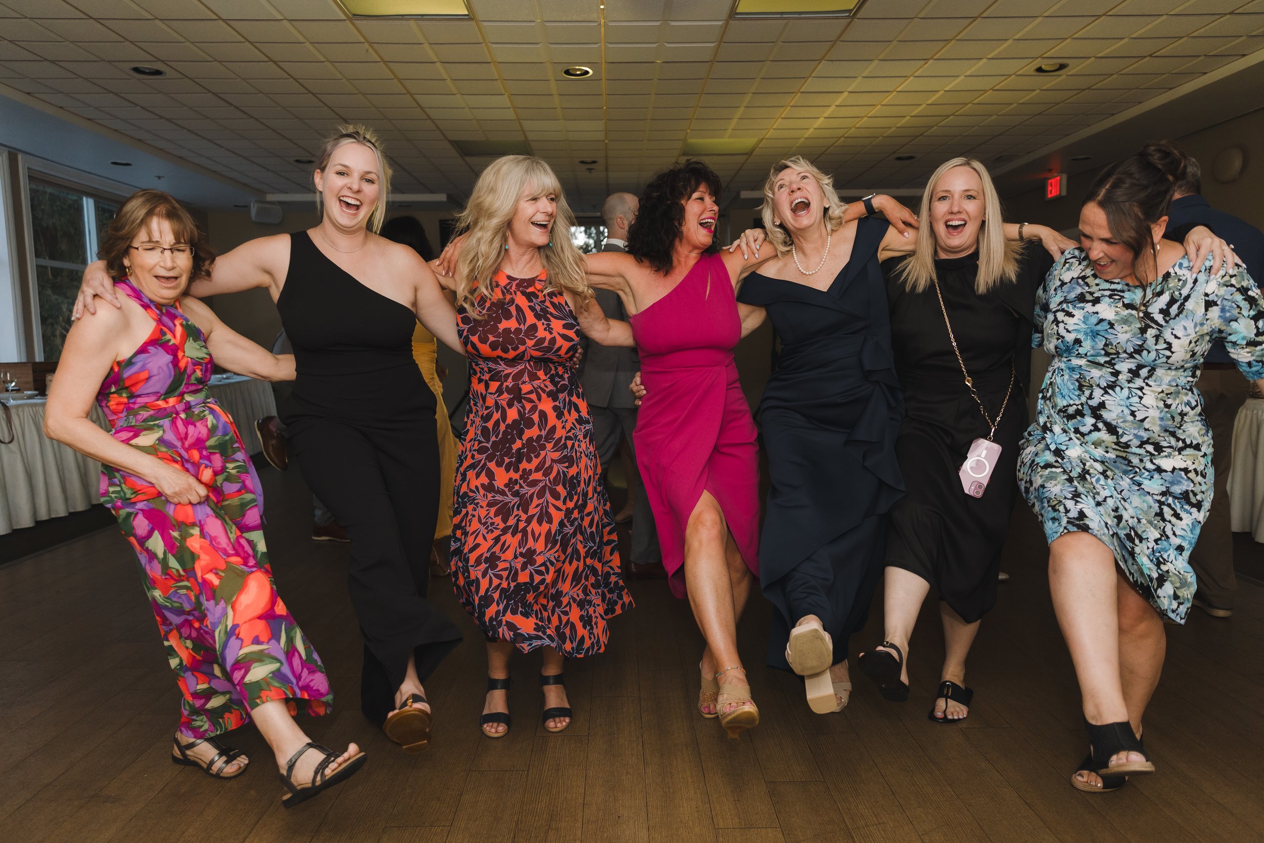 Seven women in colorful dresses and black shoes dancing together at a social event, holding each other by the shoulders or waist and laughing.