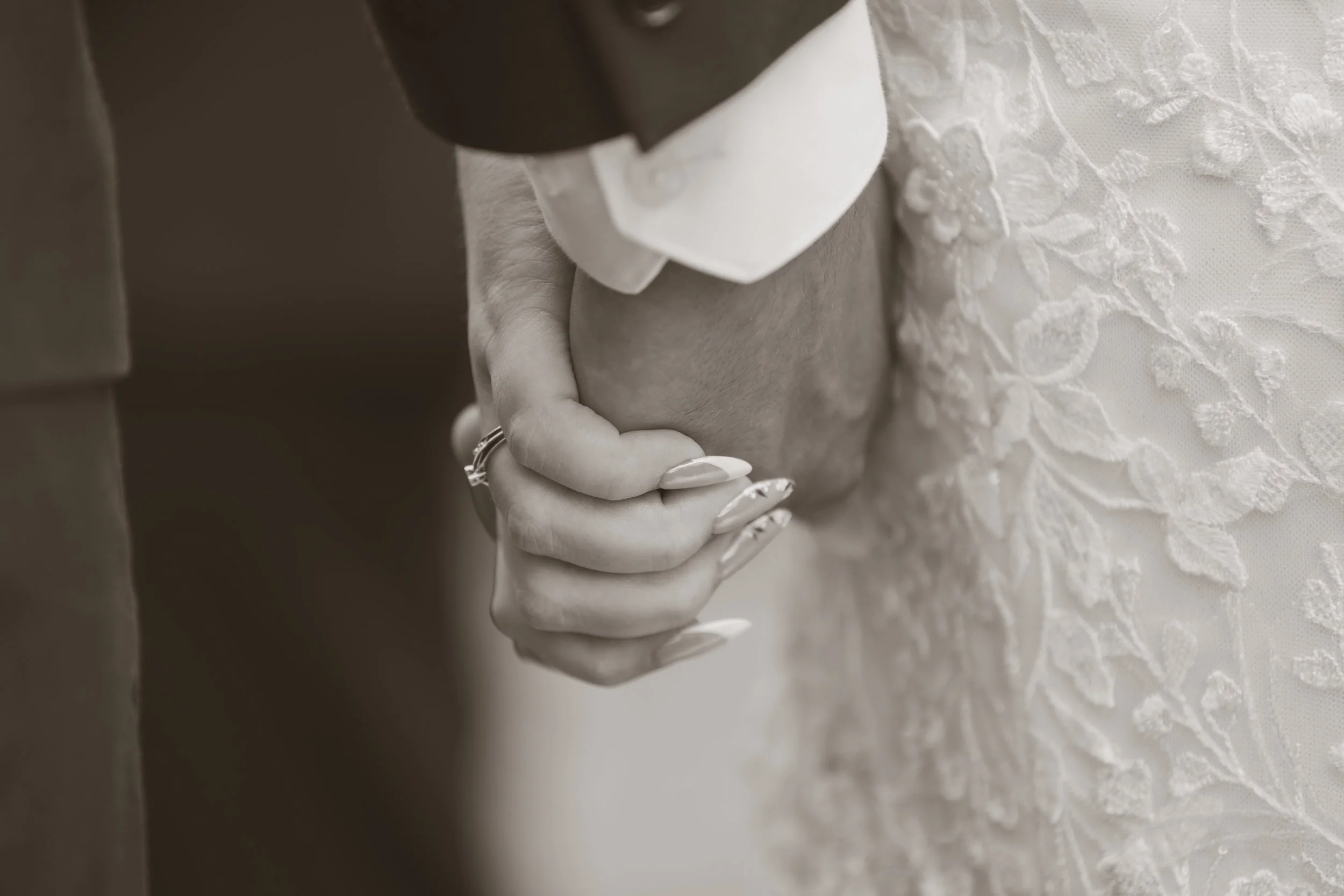 Close-up of a couple holding hands, with the woman's hand resting on top of the man's hand, both wearing wedding rings. The woman is wearing a lace wedding dress and the man has on a suit.