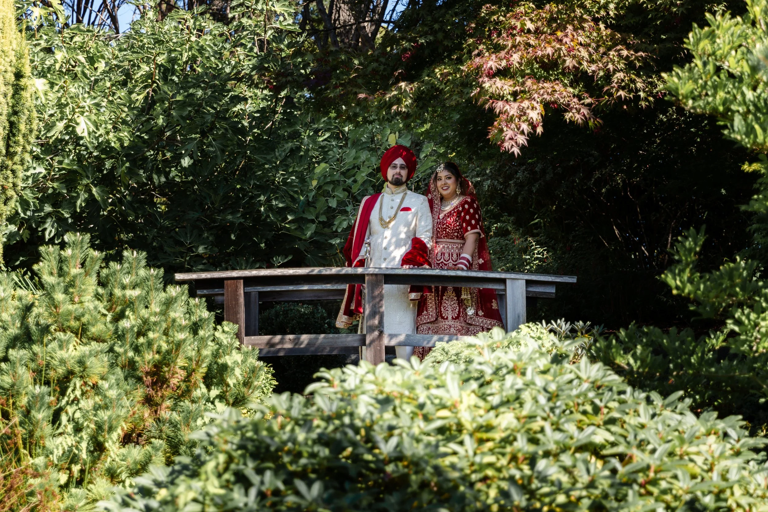 A newlywed couple dressed in traditional Indian wedding attire standing on a wooden bridge surrounded by lush green trees and bushes.