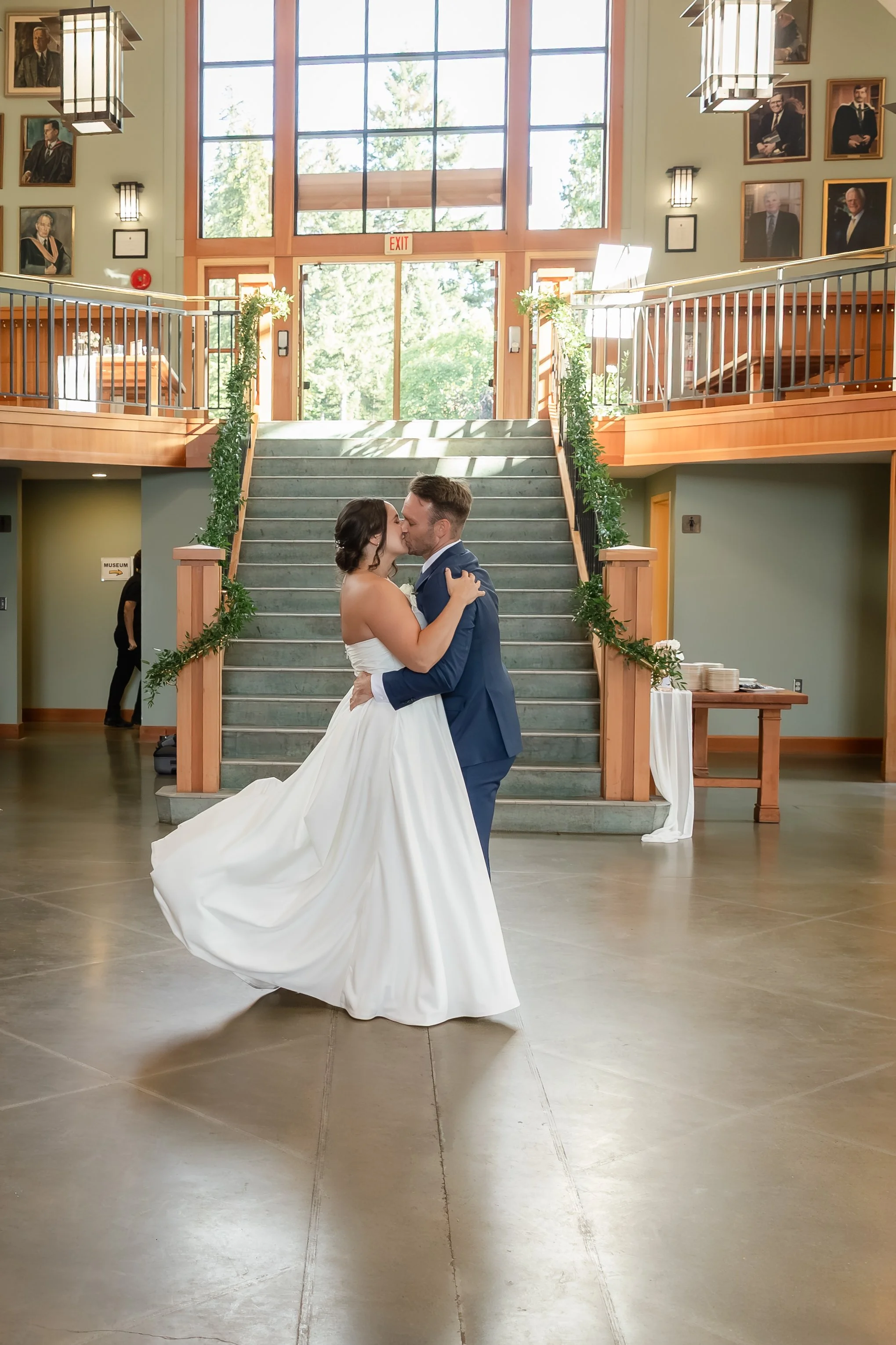 A bride and groom sharing a kiss while dancing in a large, well-lit reception hall with a staircase in the background decorated with greenery.