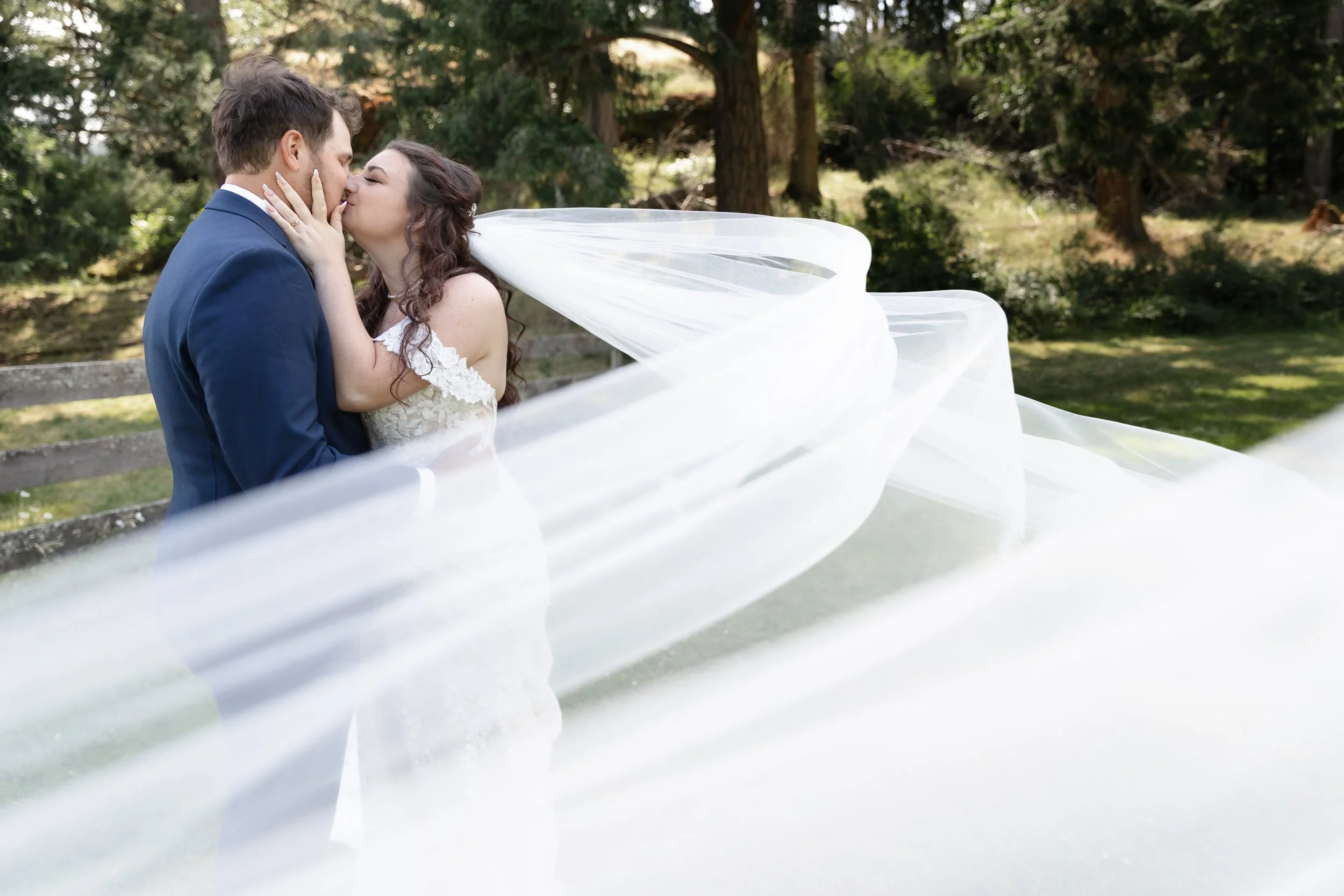 A bride and groom kissing outdoors, with the bride's flowing veil caught in motion, surrounded by trees and greenery.