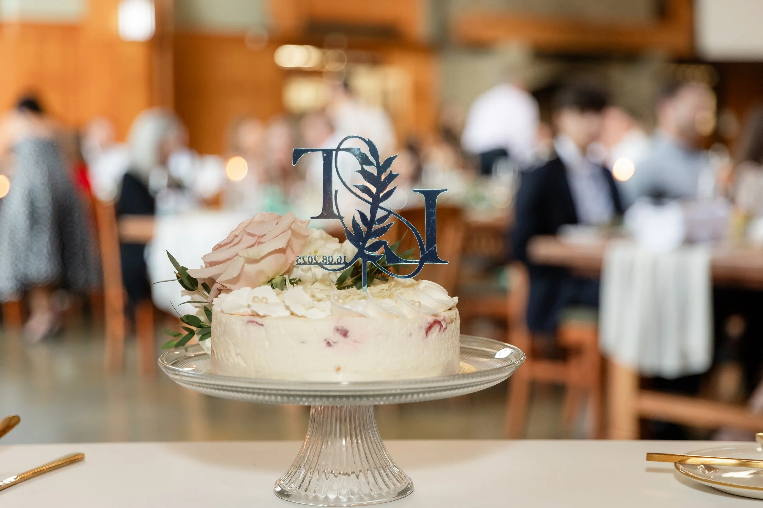A wedding cake decorated with a large pale pink rose, white icing, and a blue 'LOVE' topper, placed on a glass cake stand at a wedding reception with blurred guests and wooden decor in the background.
