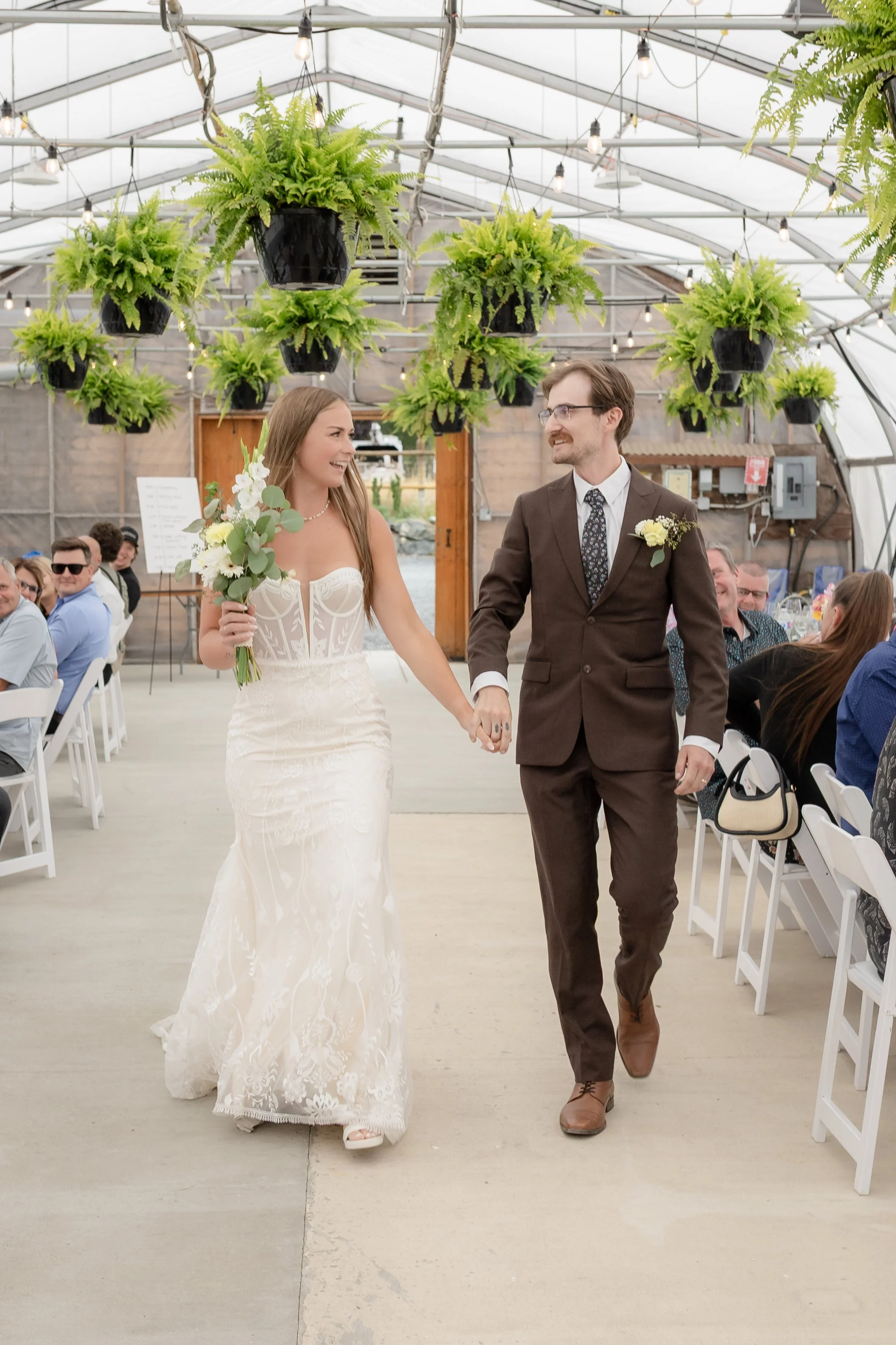 A newly married couple, the bride in a white wedding dress holding a bouquet and the groom in a brown suit, walking hand in hand down the aisle at a wedding reception in a greenhouse with hanging plants and string lights.