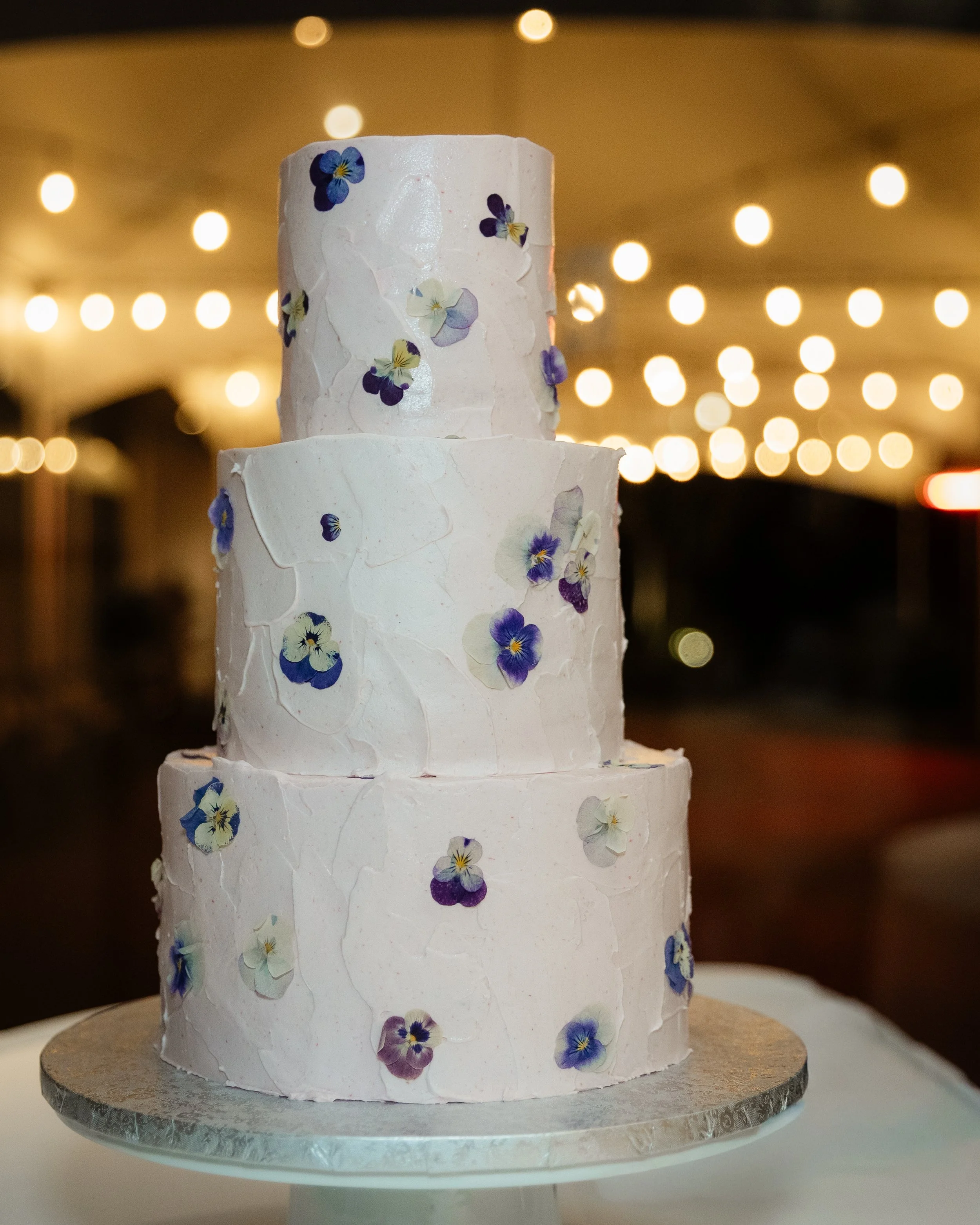 Three-tier white wedding cake decorated with edible pansy flower toppings, set on a silver cake stand with a blurred background of string lights.