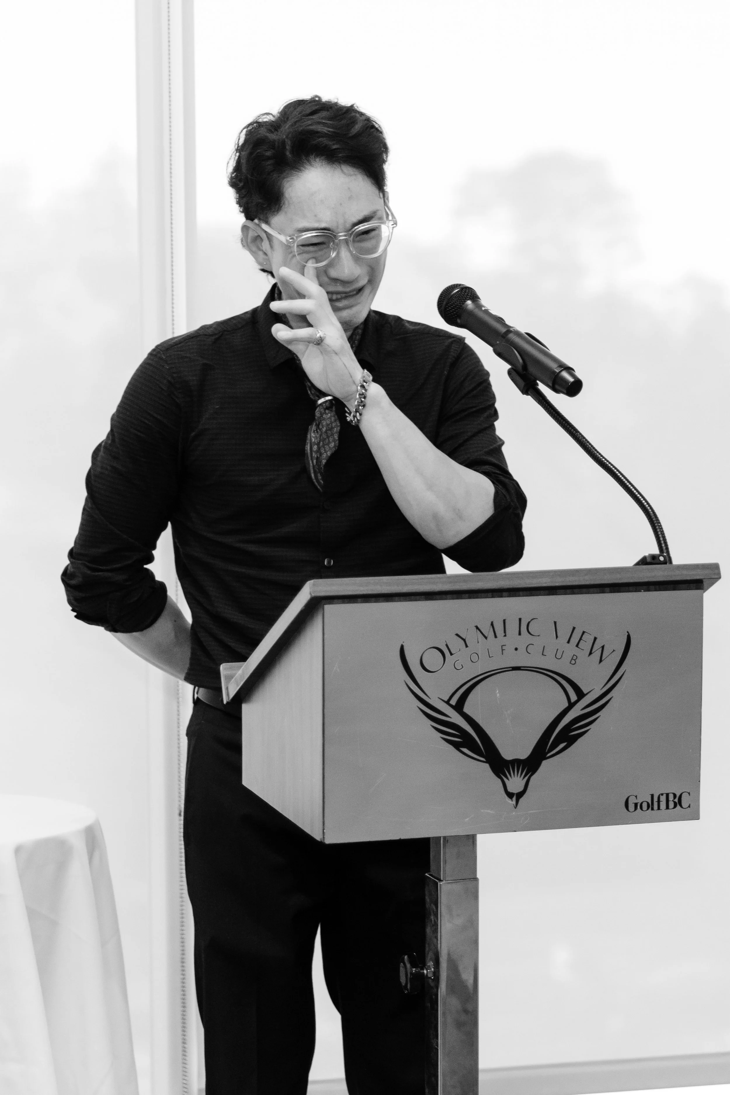 A person with short dark hair and glasses, wearing a black shirt, stands behind a podium at Olympic View Golf Club, appearing to be emotional, possibly crying.