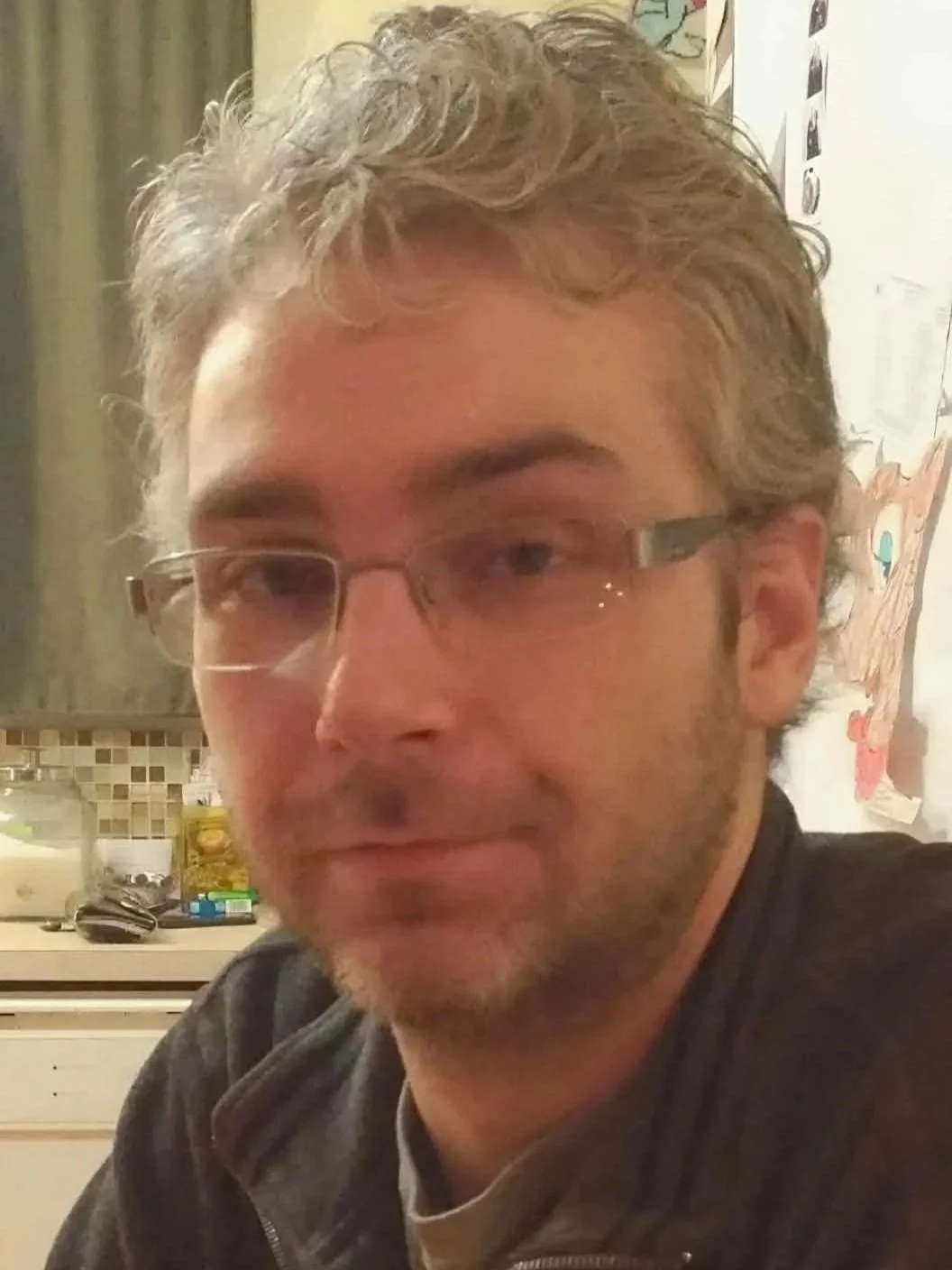 Portrait of Scott Ganley, a man with short grey-brown hair, glasses, and light facial hair. He is wearing a dark jacket and looking toward the camera. The photo is cropped into an oval shape with a white background.