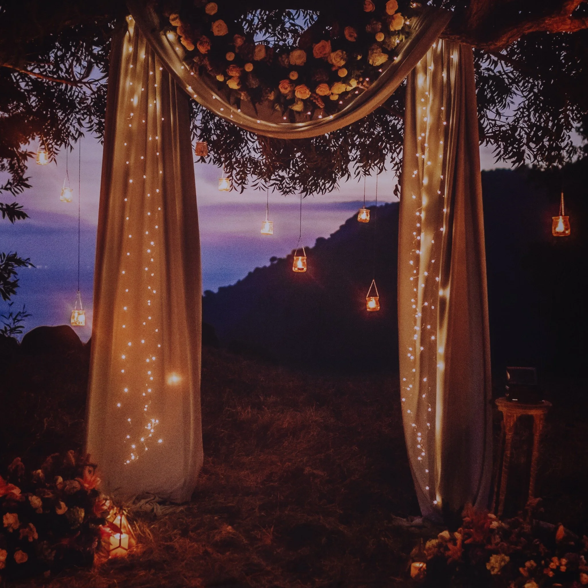 A decorated outdoor wedding arch at sunset with draped curtains, hanging lanterns, and floral arrangements, set against a backdrop of mountains and a colorful sky.