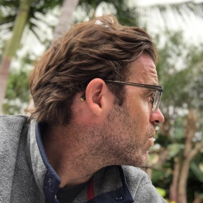 Side profile of a man with brown hair, glasses, and light beard, wearing a gray jacket, outdoors among greenery.