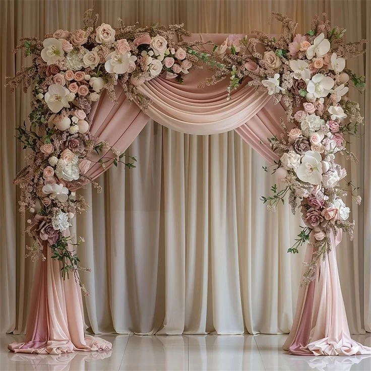 An elegant wedding arch decorated with pink and white flowers, with draped pink fabric, set against a cream-colored curtain backdrop.