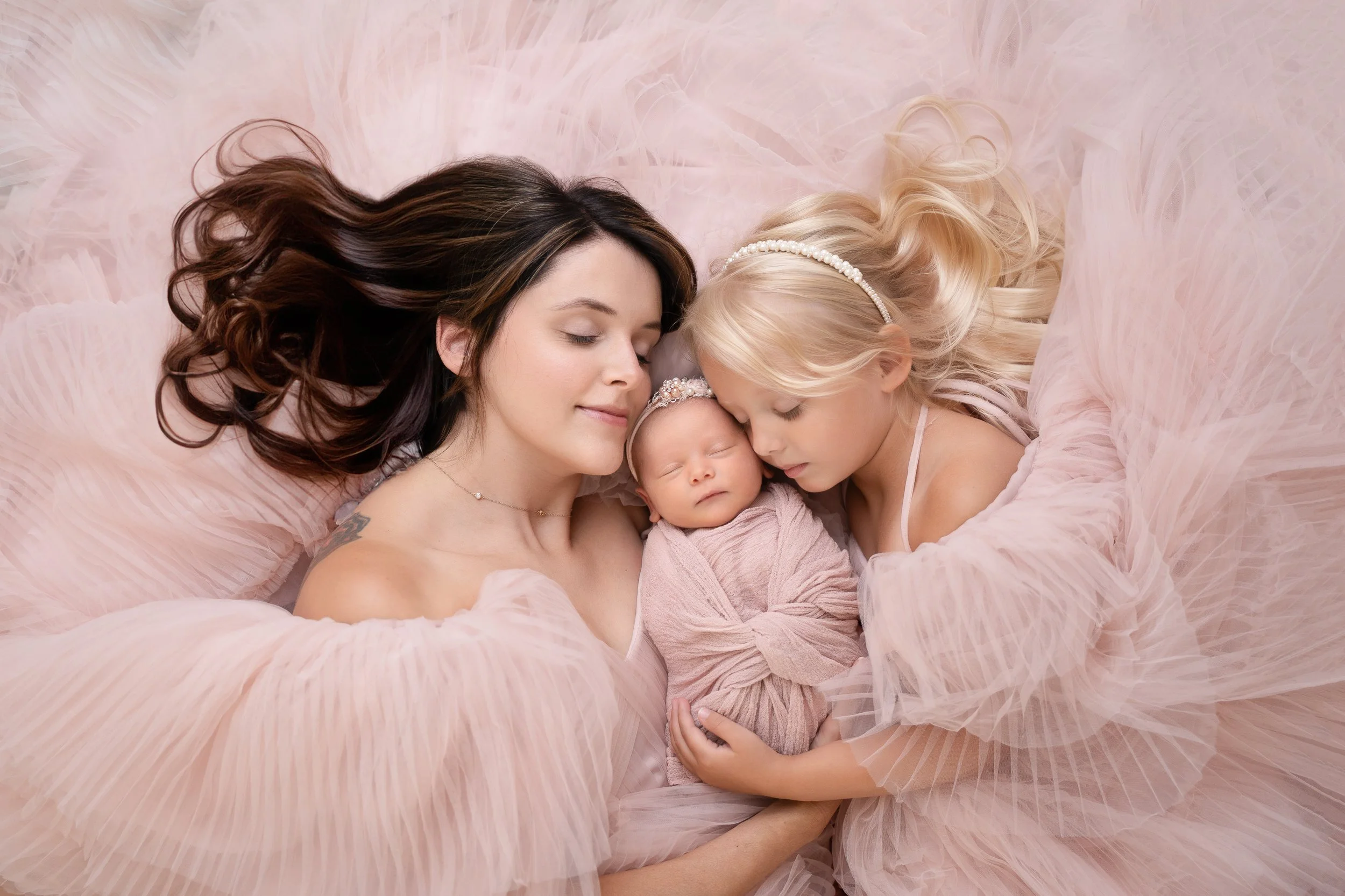 Three women, two young girls and a baby, lying close together on a soft, pink feathered blanket, sleeping peacefully. The baby, swaddled in pink, is in the center with the two girls on either side, all dressed in soft, light clothing and wearing elegant hair accessories.