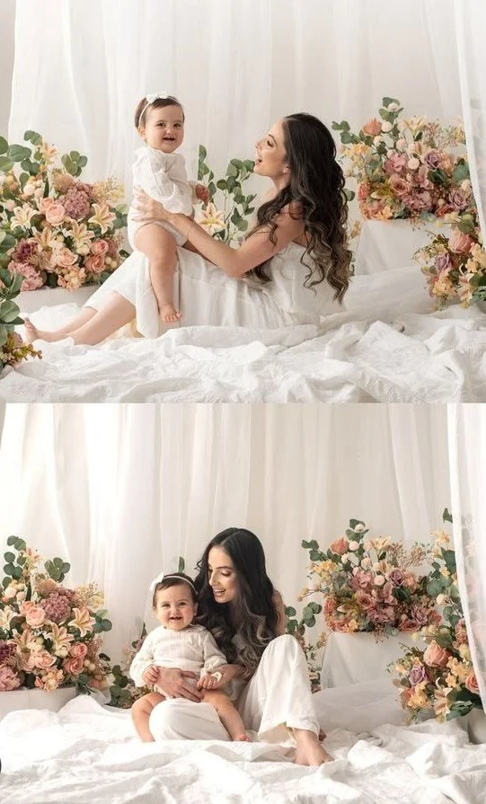 A woman with long dark hair and a little girl with a white hairband and white dress sitting and playing on a bed surrounded by pink and peach flowers with green foliage, white curtains in the background.