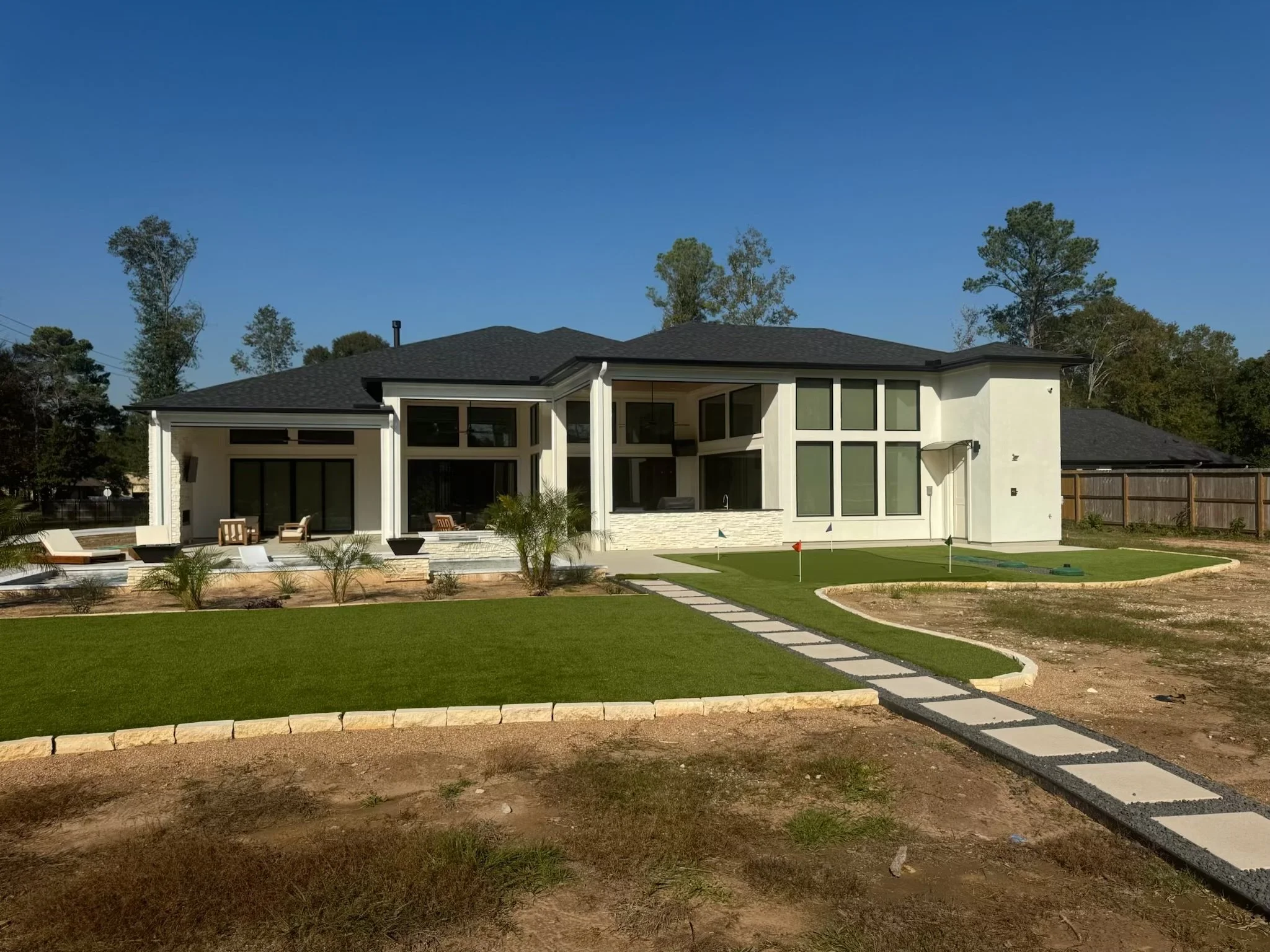 Modern house with large windows, outdoor patio furniture, a packaged putting green, and a walkway leading to the entrance, set against a backdrop of trees.