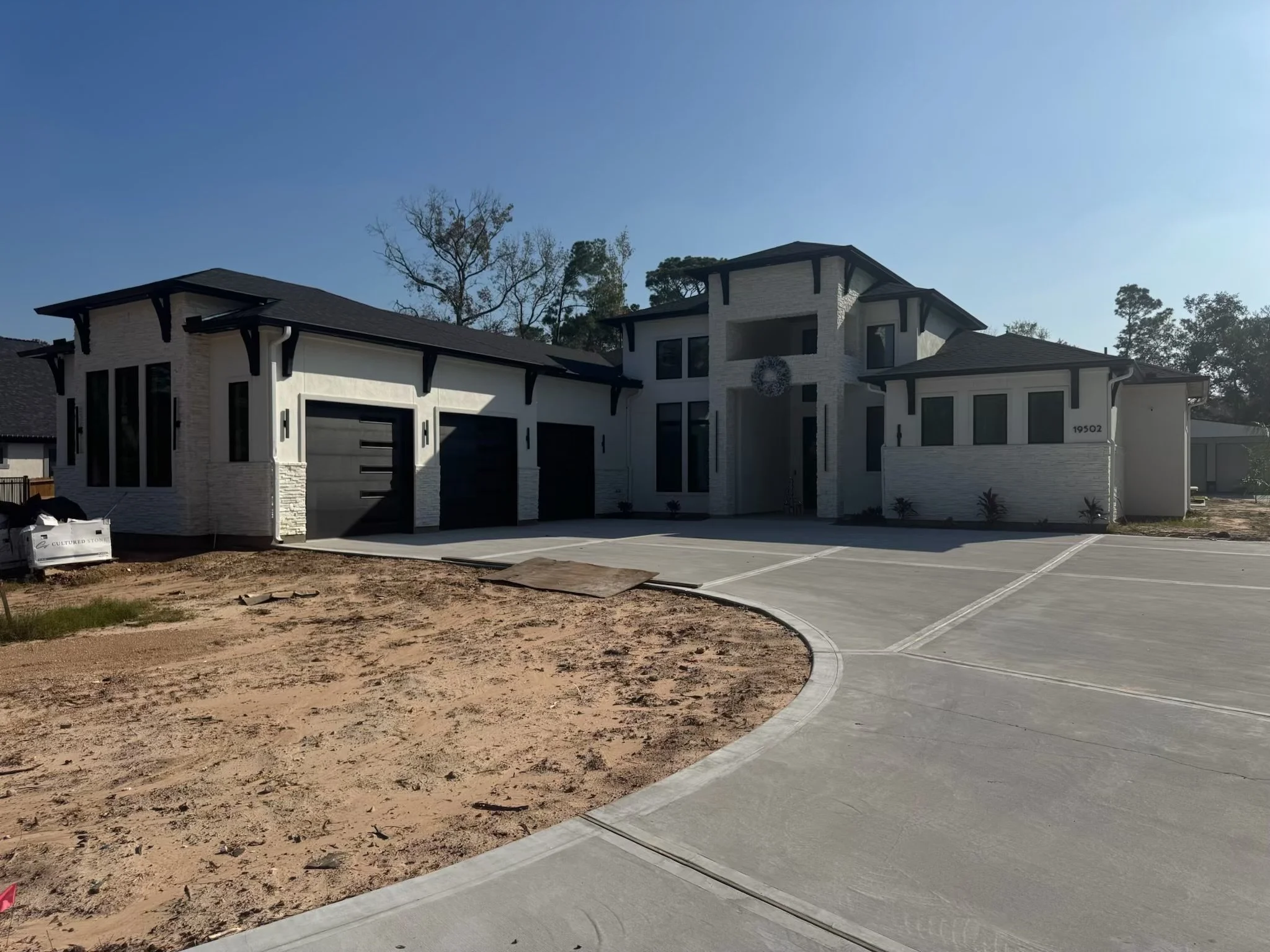 New modern house with black garage doors, white brick exterior, and black roof, surrounded by a concrete driveway and a dirt area in front.