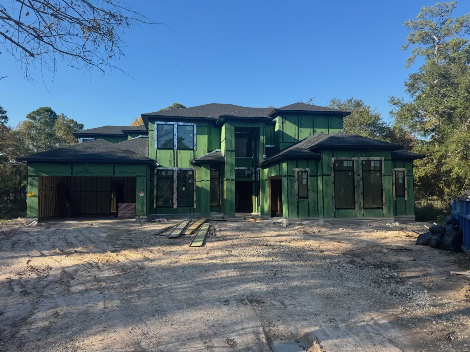 Under construction house with green sheathing and black roof, surrounded by dirt and trees.