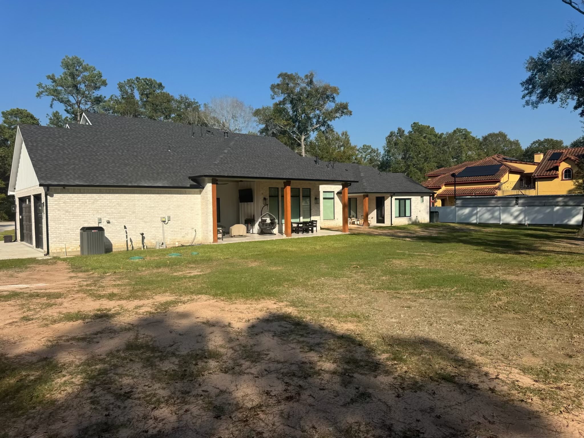 Backyard of a modern house with a covered patio, outdoor furniture, and a grassy lawn. There's a large tree line in the background and neighboring houses with red rooftops.
