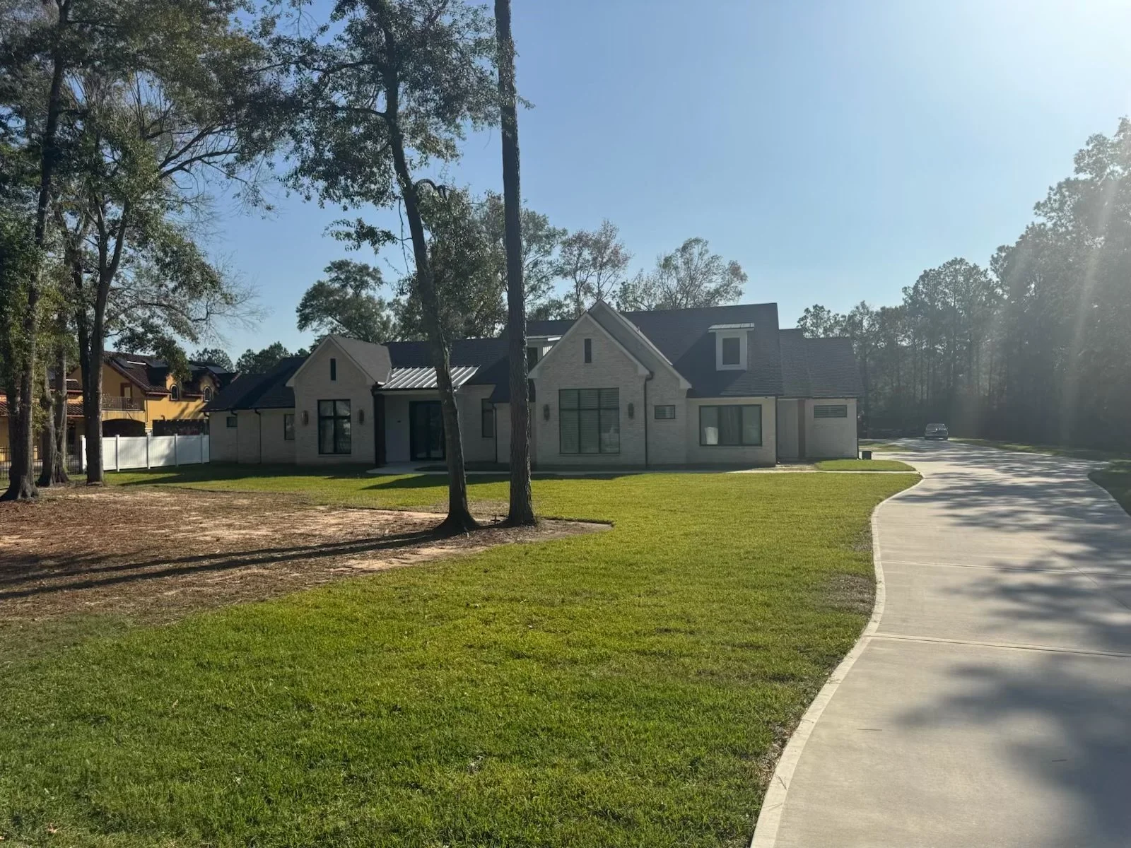A modern house with a manicured lawn, trees, and a curved sidewalk. Bright sunlight casts shadows on the greenery.