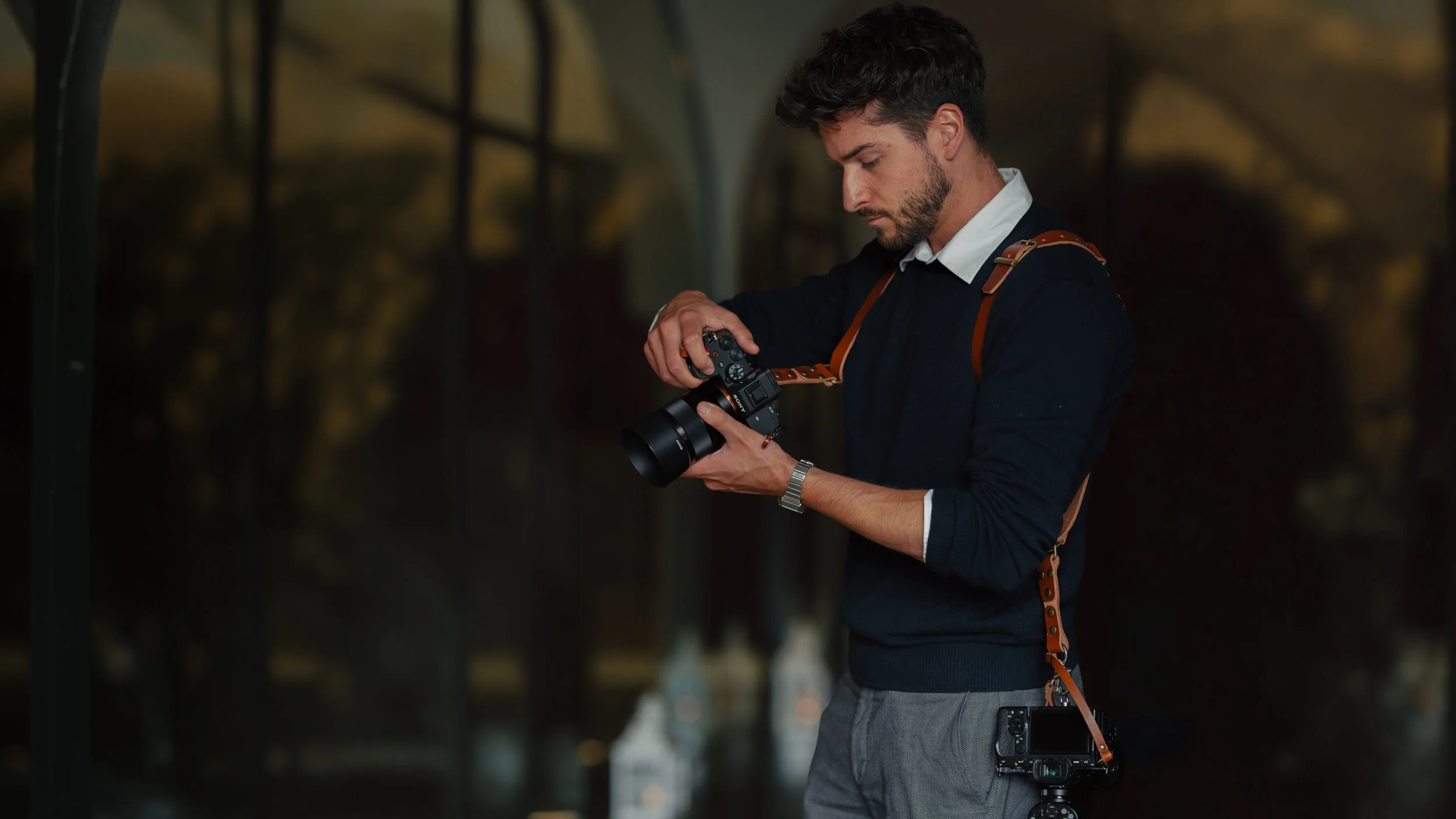 Een man met donker haar en een baard die een professionele camera instelt; hij draagt een marineblauwe trui over een wit overhemd en een grijze broek, en aan zijn heup camera nog camera , in een slecht verlichte binnenruimte.