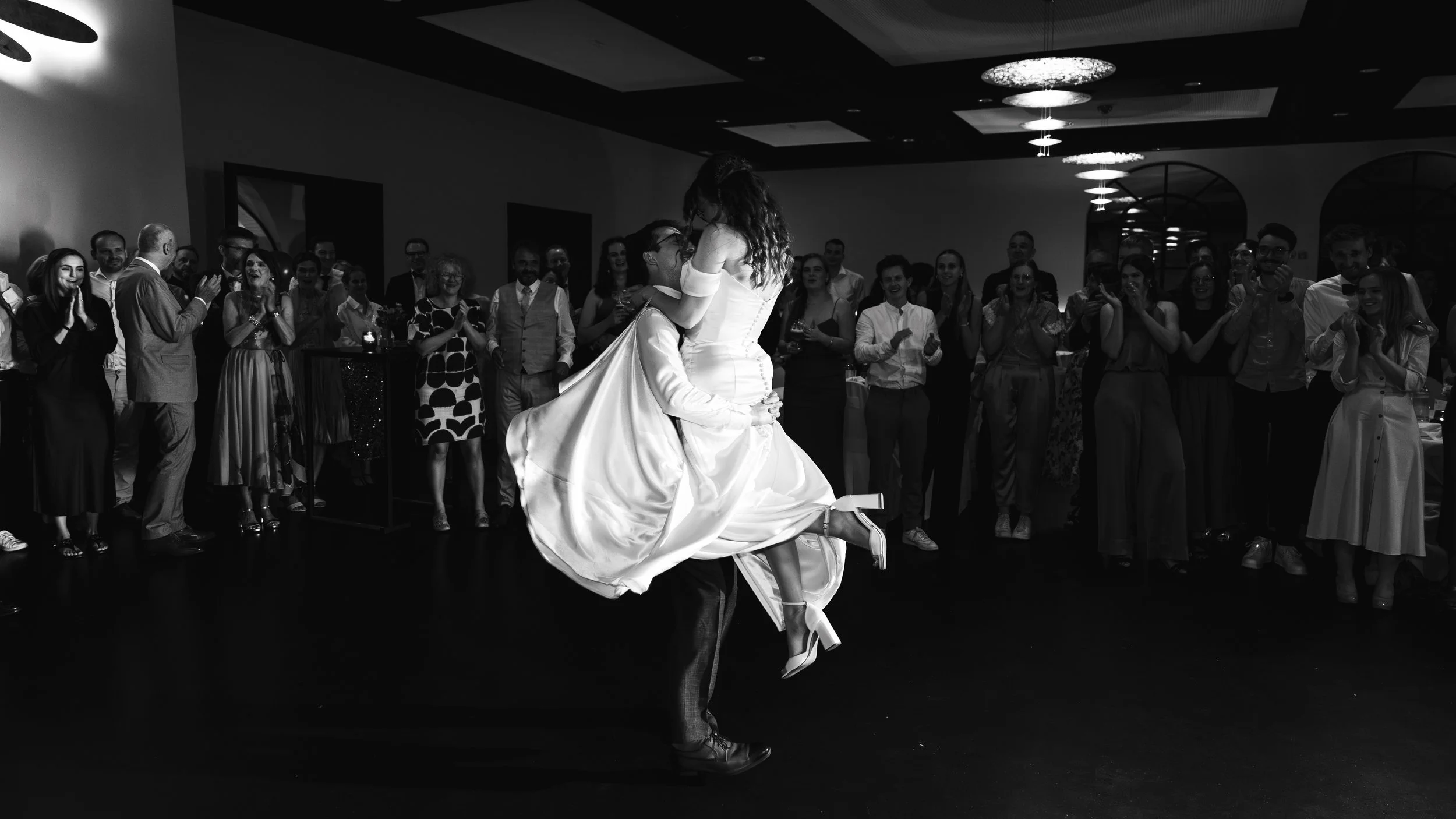 A newlywed bride being lifted in her wedding dress by the groom during a celebration with guests clapping and cheering.