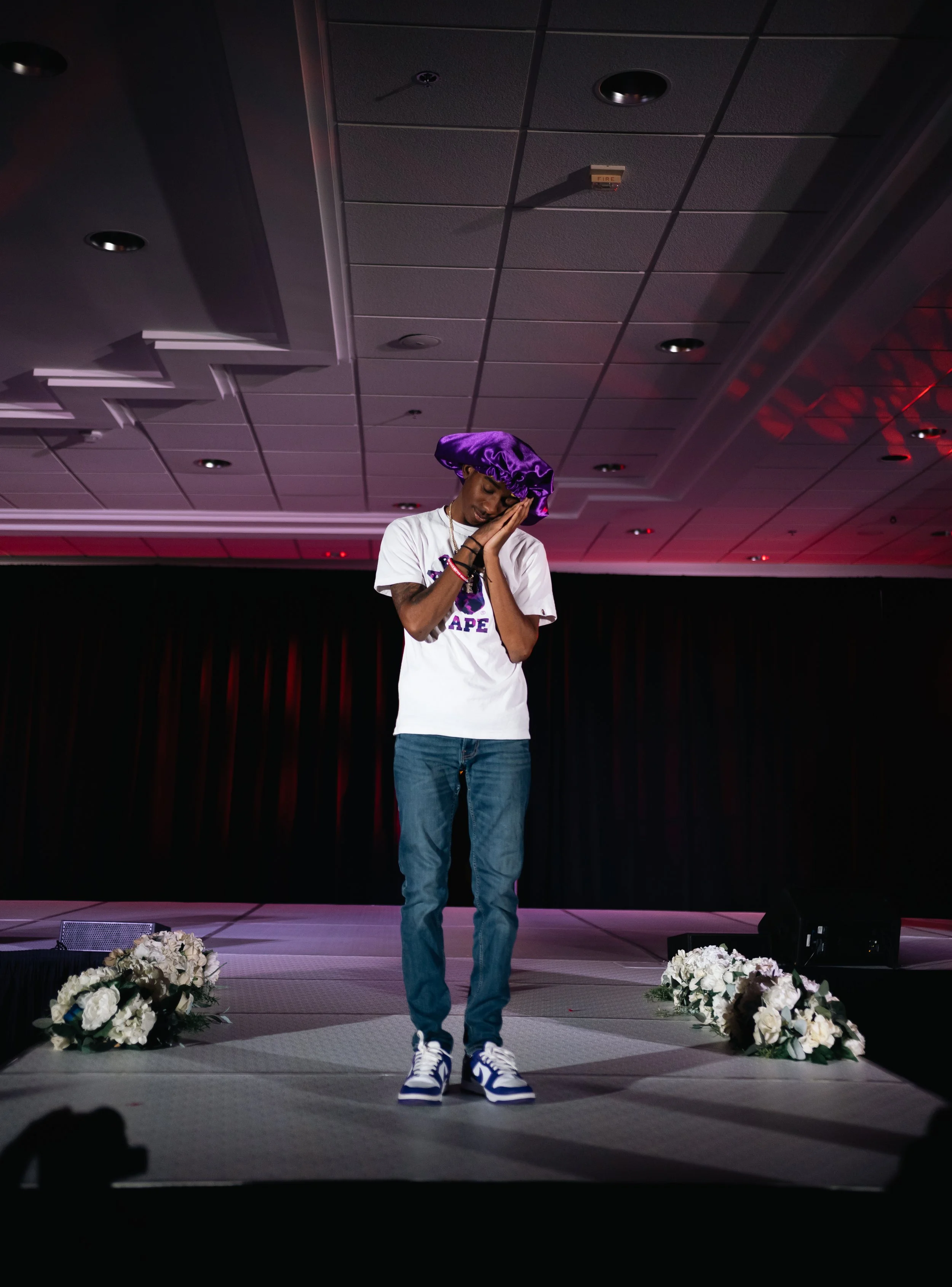 Young man standing on stage with his hands clasped near his face, wearing a purple satin bonnet, white t-shirt, jeans, and sneakers, in a ballroom with floral decorations and a black curtain backdrop.
